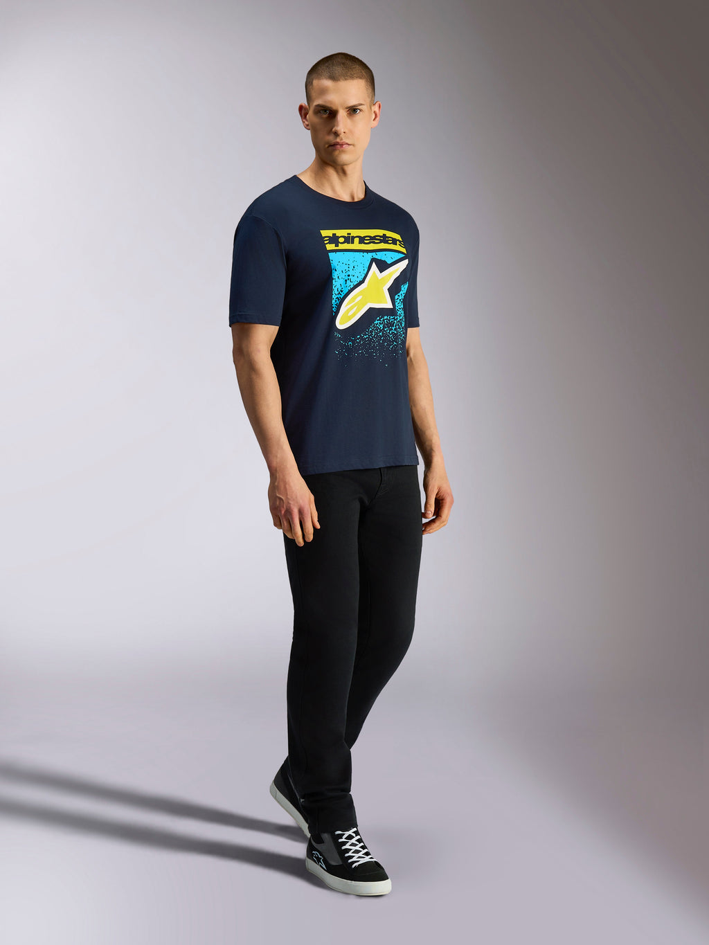 Alpinestars Noka CSF Tee, kortærmet t-shirt, marineblå, gult Astars-logo med blå MX-gradueringsgrafik, mandlig model iført marineblå t-shirt med sorte bukser og sneakers, afslappet motorsport streetwear
