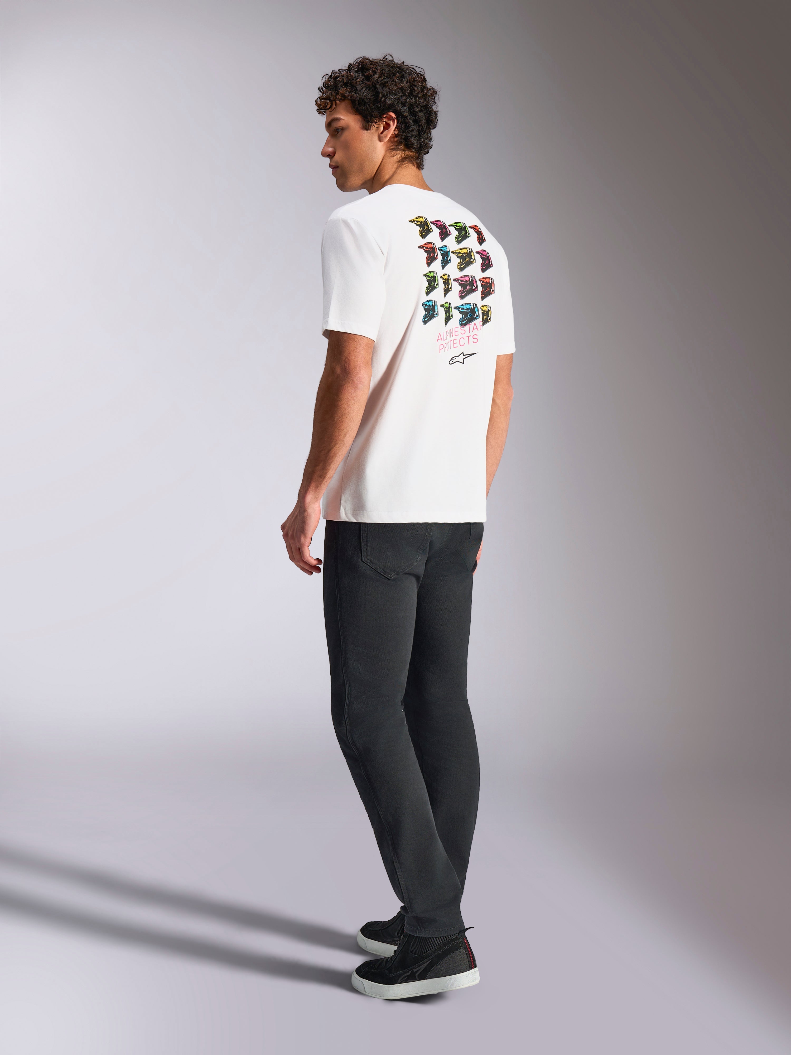 Alpinestars Pop Helmet CSF Tee, kortærmet T-shirt, hvid, set bagfra på model med farverig M10 hjelm-grafik, kombineret med mørke bukser og sorte sneakers, afslappet motorsport-inspireret streetwear