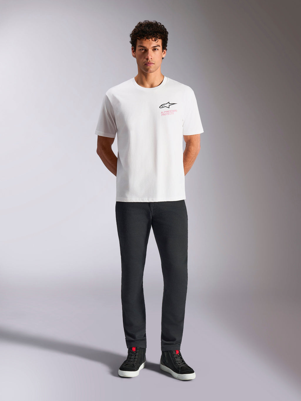Alpinestars Pop Helmet CSF Tee, kortærmet T-shirt, hvid, bæres af en model med sorte bukser og sorte high-top sneakers, Astars-logo på fronten med lyserød 'Alpinestars Protects' tekst, casual motorsport streetwear