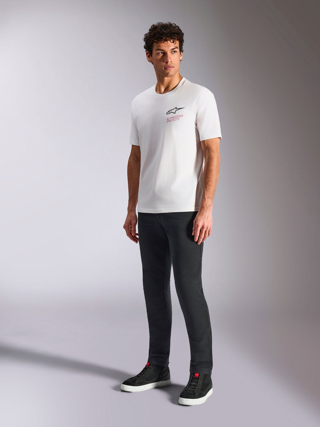 Alpinestars Pop Helmet CSF Tee, kortærmet T-shirt, hvid, med Astars-logo og rød tekst på fronten, båret af en mandlig model med mørke bukser og sorte sneakers, afslappet motorsport streetwear