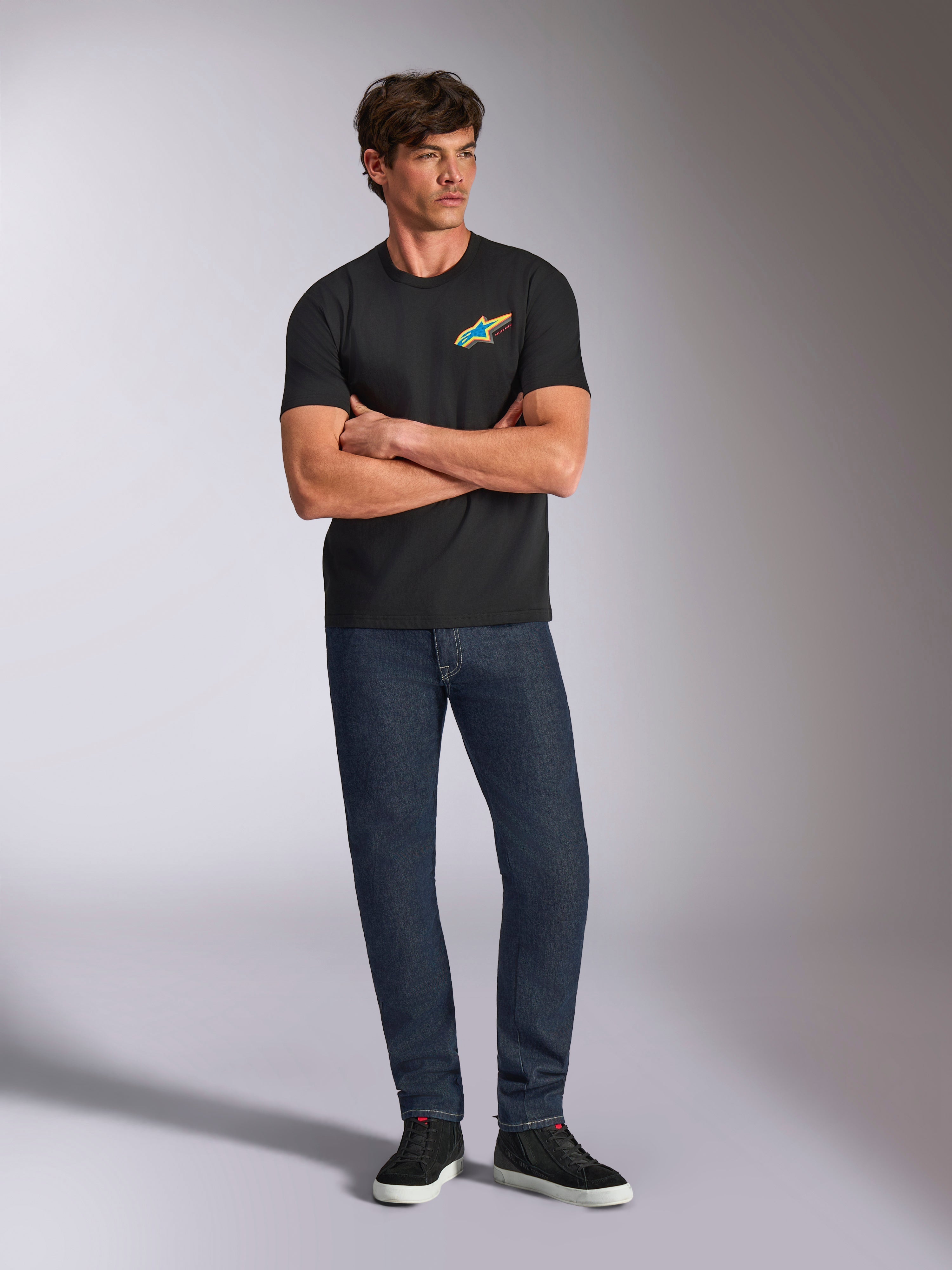 Alpinestars Angler CSF Tee, Short Sleeve T-Shirt, Black, Vibrant Multi-Color Astars Logo on Chest, worn by model with arms crossed, dark denim jeans and black sneakers, casual motorsport streetwear