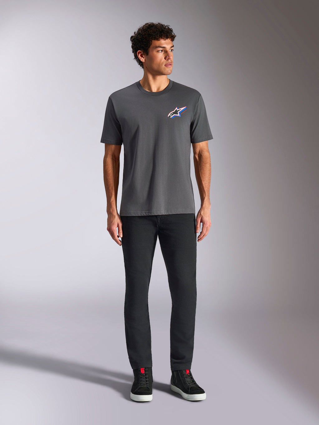 Alpinestars Angler CSF Tee, kortærmet t-shirt, koksgrå, med et livligt flerfarvet Astars-logo på brystet, bæres af en model med sorte bukser og sneakers, afslappet motorsport-streetwear