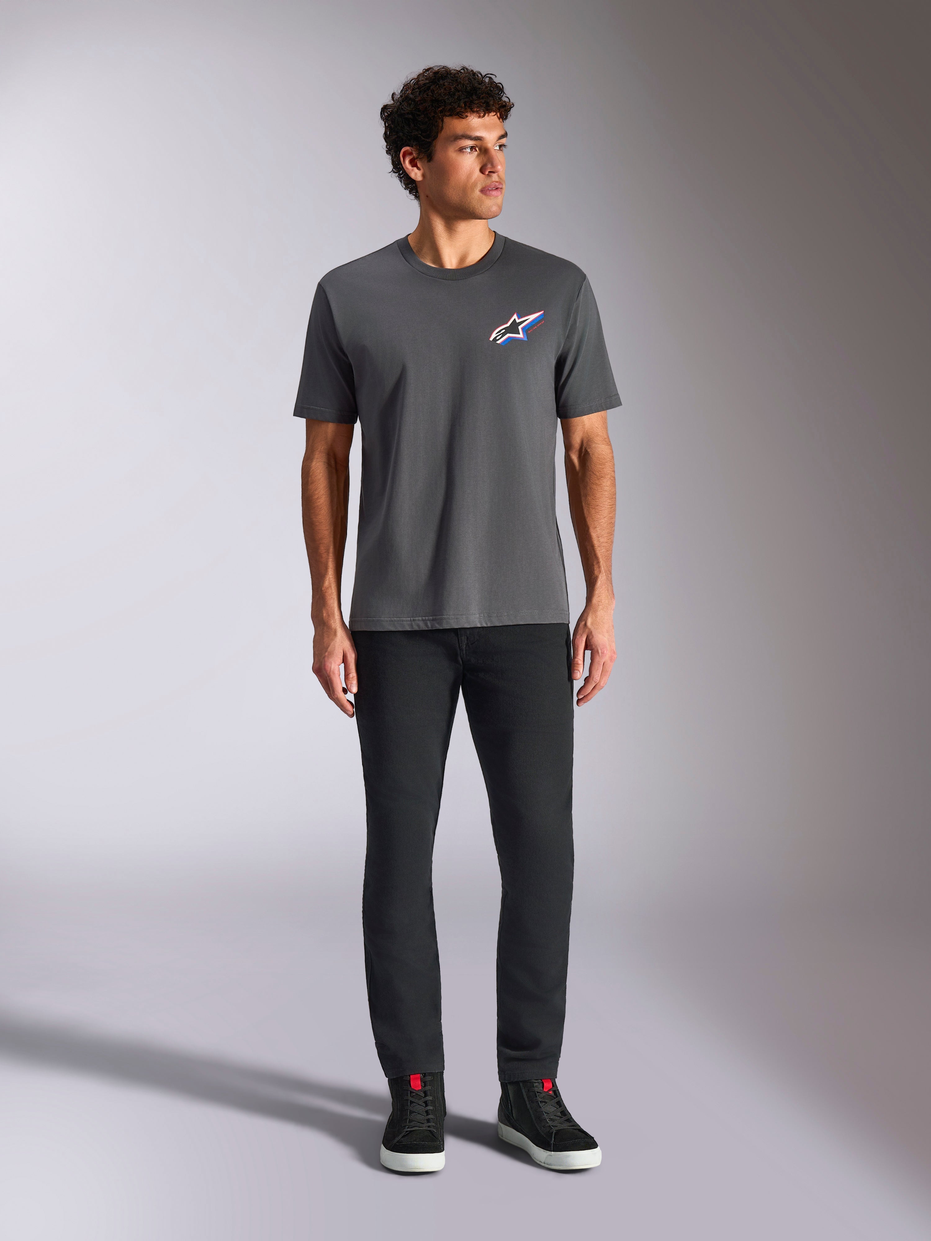 Alpinestars Angler CSF Tee, kortærmet t-shirt, koksgrå, med et livligt flerfarvet Astars-logo på brystet, bæres af en model med sorte bukser og sneakers, afslappet motorsport-streetwear