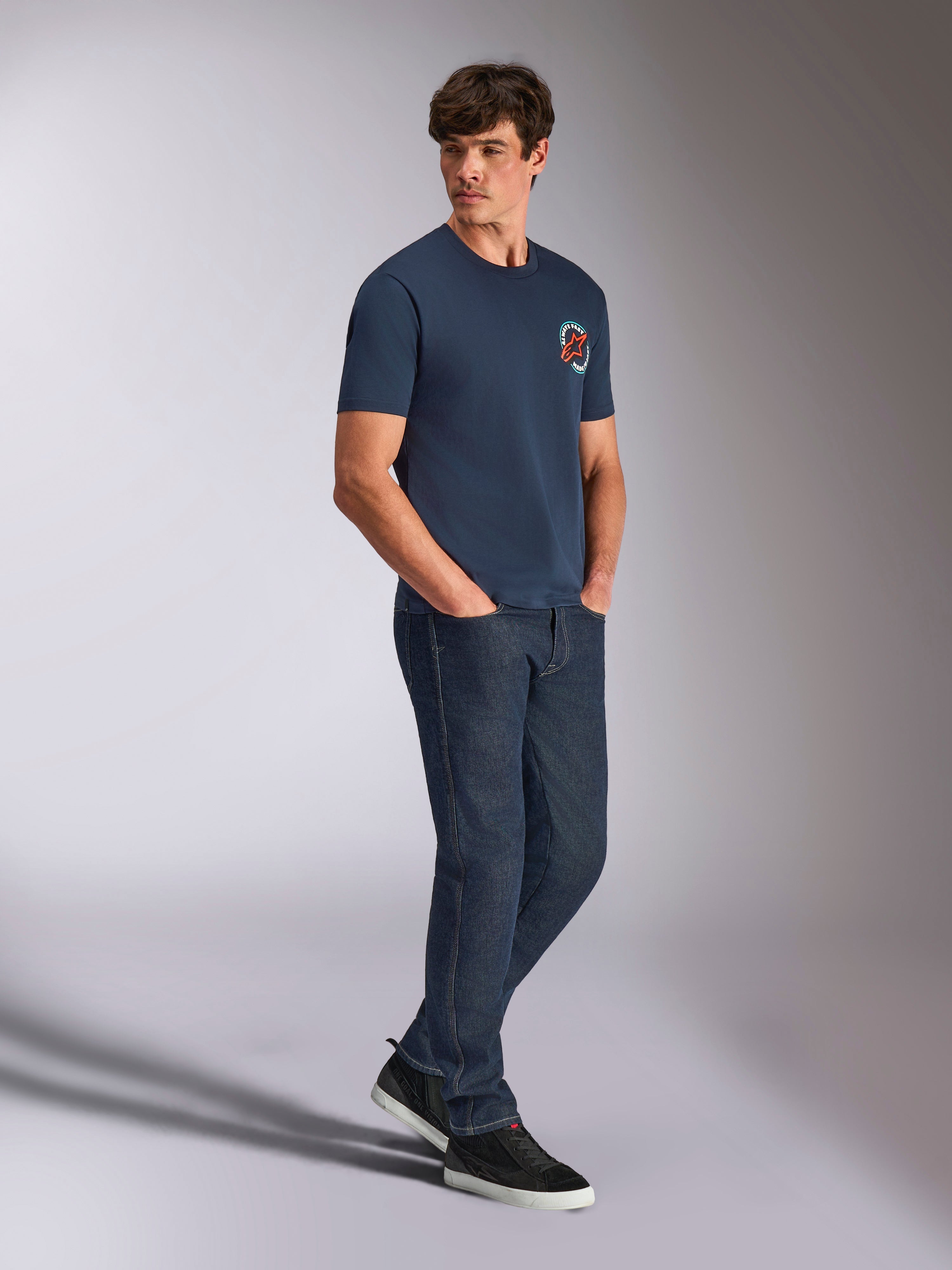 Alpinestars Aorbit CSF Tee, kortærmet T-shirt, navy, med cirkulært orange og hvidt Astars-logo med Always Fast Made to Last-grafik, båret af en mand med mørke denimjeans og sorte sneakers, afslappet motorsport-inspireret streetwear