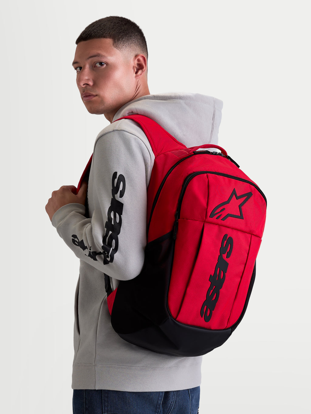 Alpinestars GFX Boss Backpack, 20L Urban Sportswear Daypack, Red and Black, shown on model wearing a grey hoodie. Featuring durable 600D polyester construction, raised silicone Astars graphic branding, and internal laptop sleeve. Ideal for daily commuting and everyday carry.