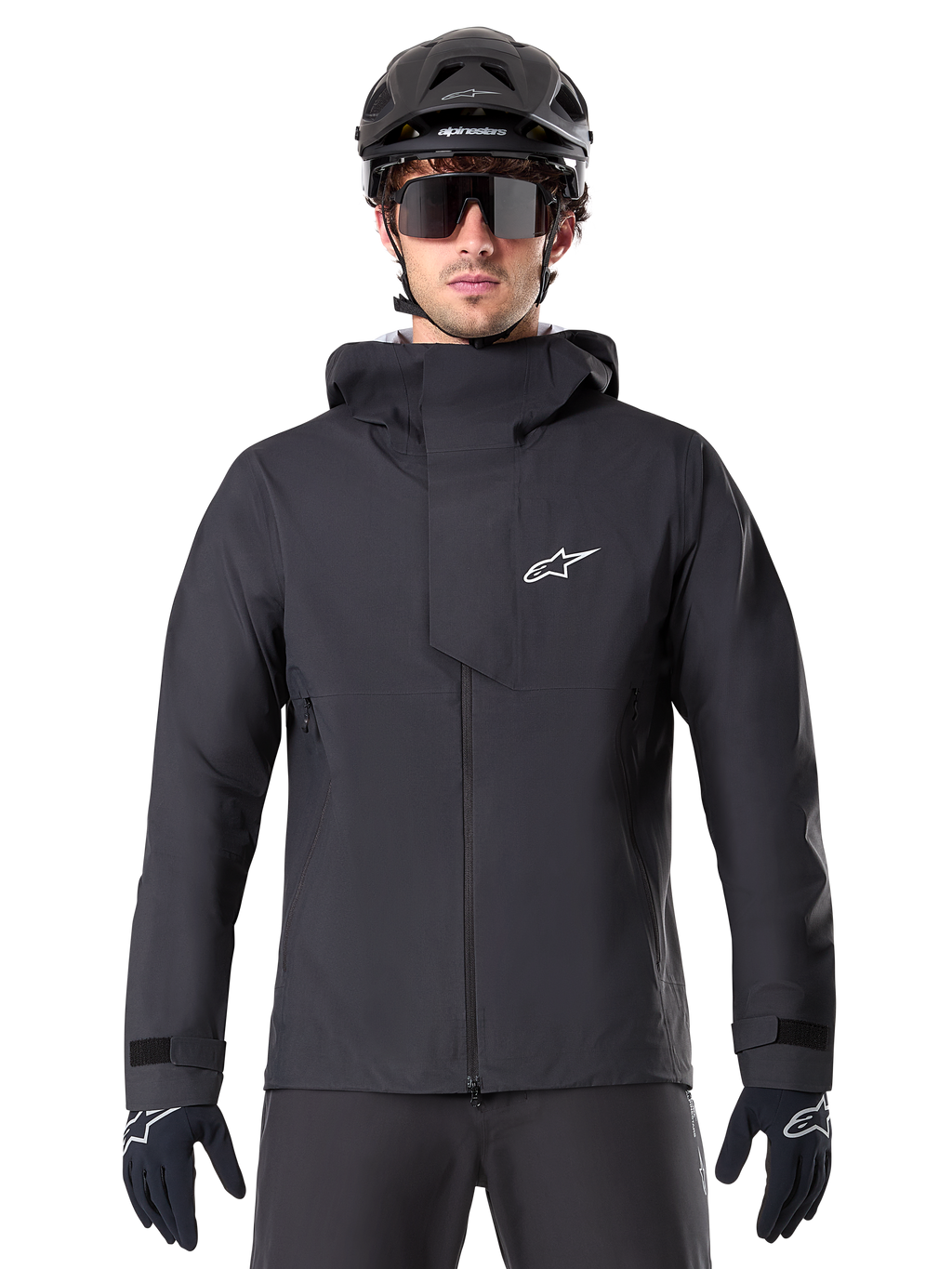 Alpinestars A-Dura Elite vandtæt jakke, sort, mountainbike-jakke, design med høj krave og hætte, lynlås foran med beskyttende brystklap og hvidt logo, vist med sort hjelm og cykelhandsker, teknisk vejrbeskyttelse til trail-kørsel