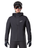 Alpinestars A-Dura Elite vandtæt jakke, sort, mountainbike-jakke, design med høj krave og hætte, lynlås foran med beskyttende brystklap og hvidt logo, vist med sort hjelm og cykelhandsker, teknisk vejrbeskyttelse til trail-kørsel