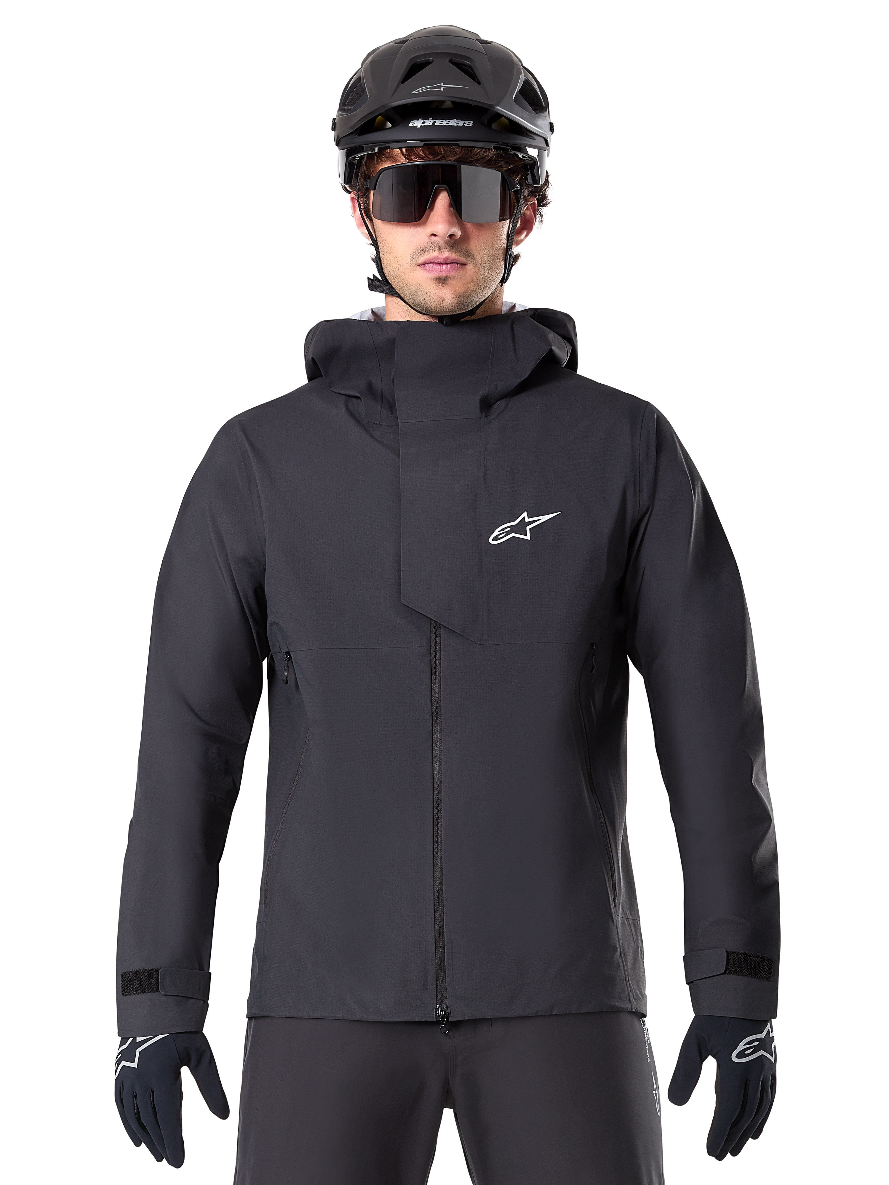 Alpinestars A-Dura Elite vandtæt jakke, sort, mountainbike-jakke, design med høj krave og hætte, lynlås foran med beskyttende brystklap og hvidt logo, vist med sort hjelm og cykelhandsker, teknisk vejrbeskyttelse til trail-kørsel