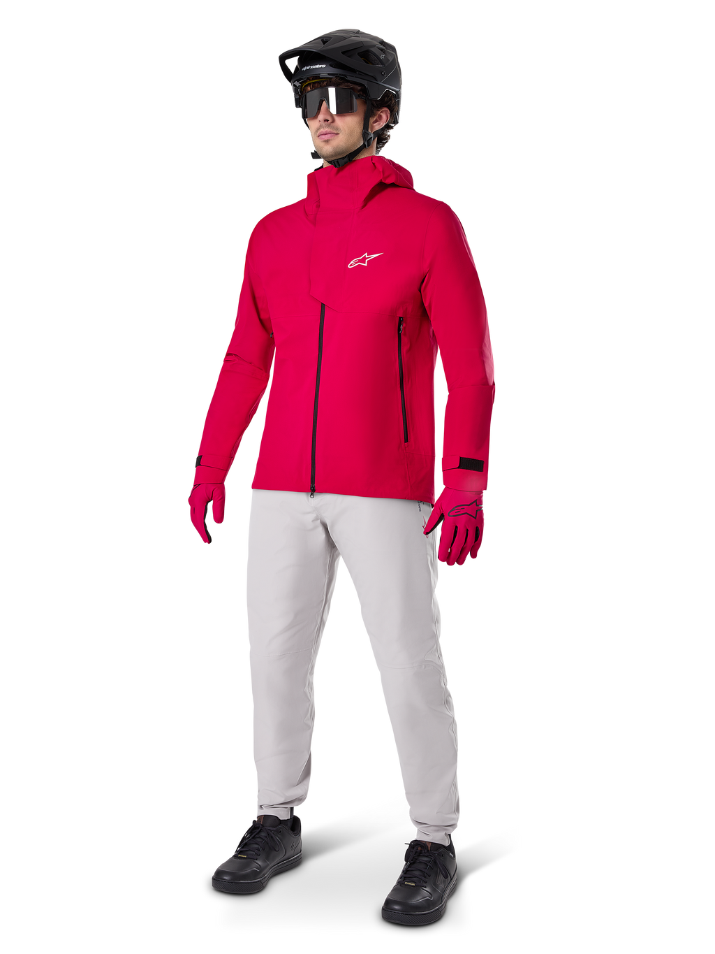 Mandlig cyklist iført Alpinestars A-Dura Elite vandtæt jakke i Red Fluo, kombineret med lysegrå tekniske bukser, røde handsker, sort MTB-hjelm med goggles og sorte sko, komplet højtydende outfit til mountainbiking
