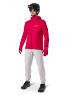 Mandlig cyklist iført Alpinestars A-Dura Elite vandtæt jakke i Red Fluo, kombineret med lysegrå tekniske bukser, røde handsker, sort MTB-hjelm med goggles og sorte sko, komplet højtydende outfit til mountainbiking