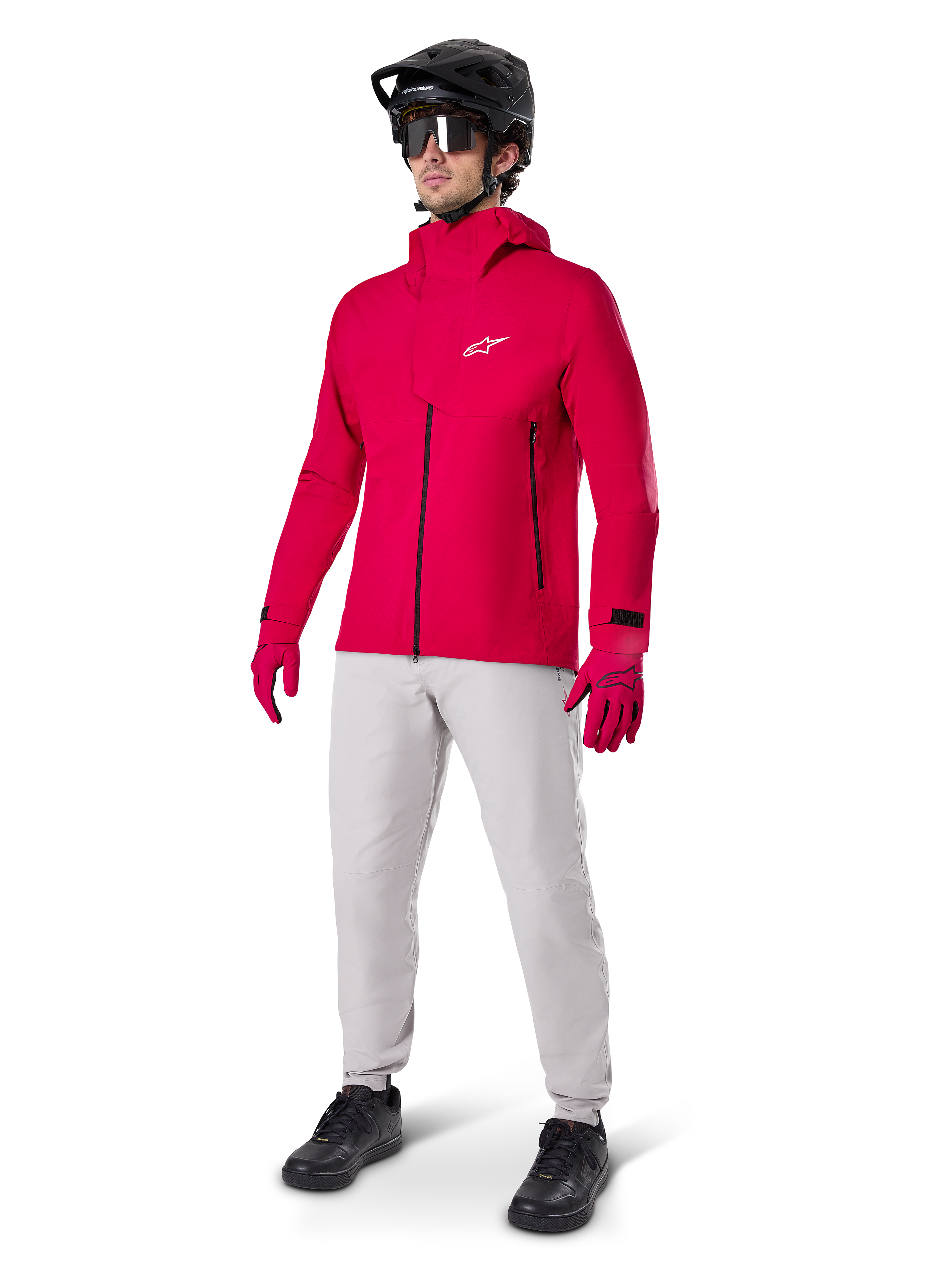 Mandlig cyklist iført Alpinestars A-Dura Elite vandtæt jakke i Red Fluo, kombineret med lysegrå tekniske bukser, røde handsker, sort MTB-hjelm med goggles og sorte sko, komplet højtydende outfit til mountainbiking