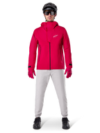 Alpinestars A-Dura Elite vandtæt jakke, Red Fluo, teknisk mountainbike-jakke båret med en sort MTB-hjelm, solbriller, matchende røde handsker og lysegrå bukser, fuldkropsvisning af cykeludstyr til alle vejrforhold