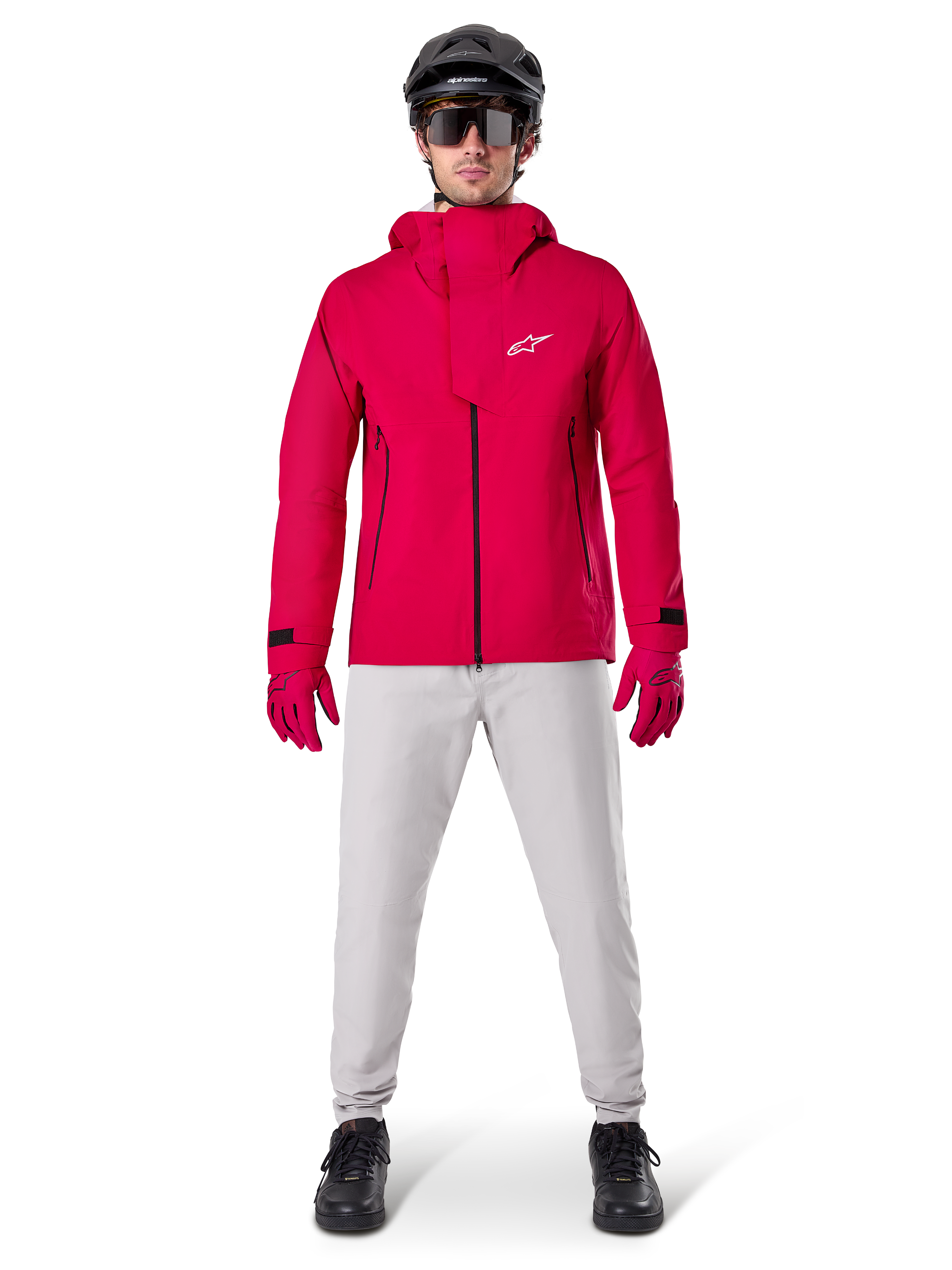 Alpinestars A-Dura Elite vandtæt jakke, Red Fluo, teknisk mountainbike-jakke båret med en sort MTB-hjelm, solbriller, matchende røde handsker og lysegrå bukser, fuldkropsvisning af cykeludstyr til alle vejrforhold