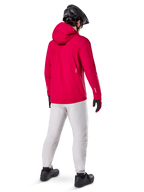 Alpinestars A-Dura Elite vandtæt jakke, Red Fluo, set bagfra i trekvart profil, båret med lysegrå bukser og sort MTB-hjelm, har hætte og reflekterende detaljer, højtydende cykelregnjakke til trail-kørsel