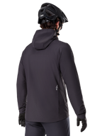 Alpinestars A-Dura Thermal Jacket, sort, set bagfra, båret med en sort mountainbikehjelm og handsker, har et lille hvidt Astars-logo nederst på ryggen, letvægts isoleret cykeljakke