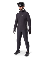 Alpinestars A-Dura Thermal jakke i sort, båret af en mandlig rytter med komplet mountainbike-udstyr inklusive sort hjelm, solbriller og cykelbukser, let isoleret jakke med hætte til cykelperformance i koldt vejr.