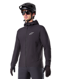 Alpinestars A-Dura Thermal Jacket, sort cykeljakke, design med hætte og lynlås foran, hvidt stjernelogo på brystet, vist med sort mountainbikehjelm og handsker, sporty performance-udstyr til terrænkørsel i koldt vejr