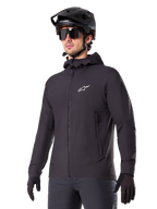 Alpinestars A-Dura Thermal Jacket, sort cykeljakke, design med hætte og lynlås foran, hvidt stjernelogo på brystet, vist med sort mountainbikehjelm og handsker, sporty performance-udstyr til terrænkørsel i koldt vejr