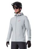 Alpinestars A-Dura Thermal Jacket, light gray insulated cycling jacket, hooded design with a small red logo, worn with a black helmet and gloves, technical mountain bike apparel for cold-weather riding