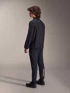 Alpinestars A-Aria Windbreaker Jacket, Black, lightweight wind-resistant cycling jacket, rear three-quarter view on model, featuring a small white logo on the back neck and elasticated hem, paired with matching black pants and shoes for a performance-driven look.