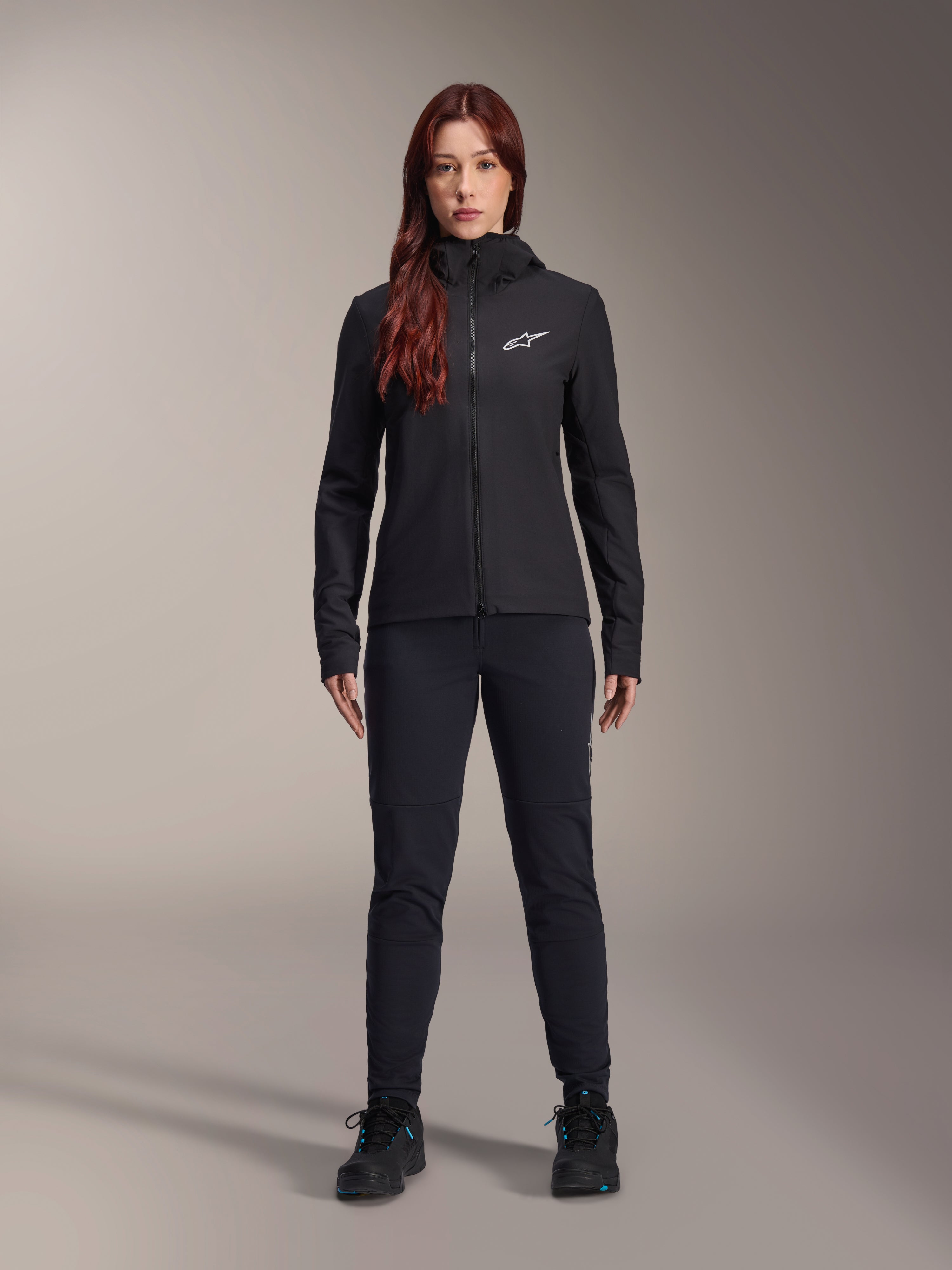 Kvinde iført Alpinestars Stella A-Dura Thermal Jacket, sort letvægtsisoleret cykeljakke med hætte og hvidt Astars-logo på brystet, kombineret med sorte bukser og sportssko