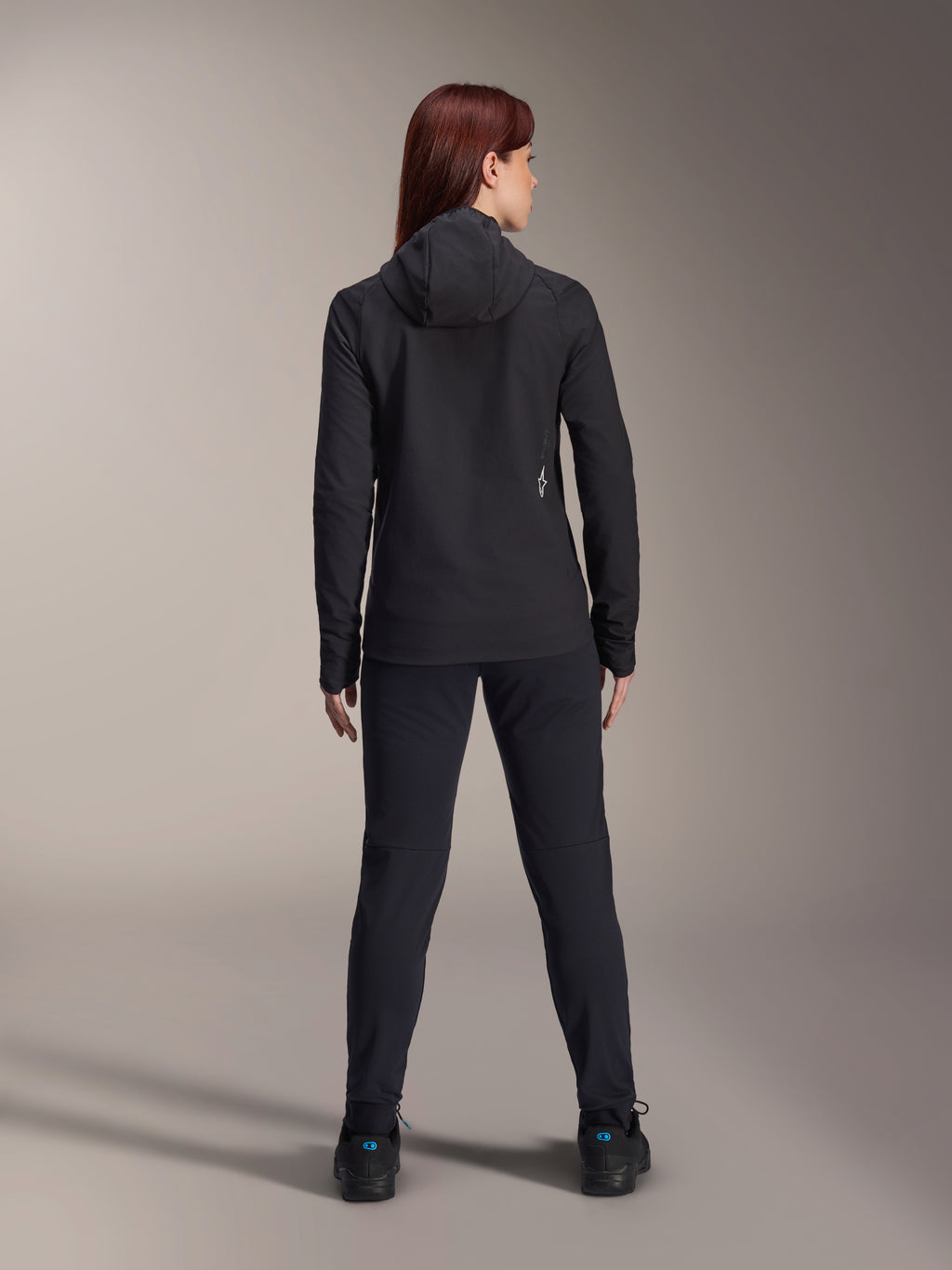 Kvinde iført Alpinestars Stella A-Dura Termojakke, set bagfra, sort isoleret cykeljakke med hætte, matchende sorte bukser og sko, lille hvidt logo nederst på ryggen