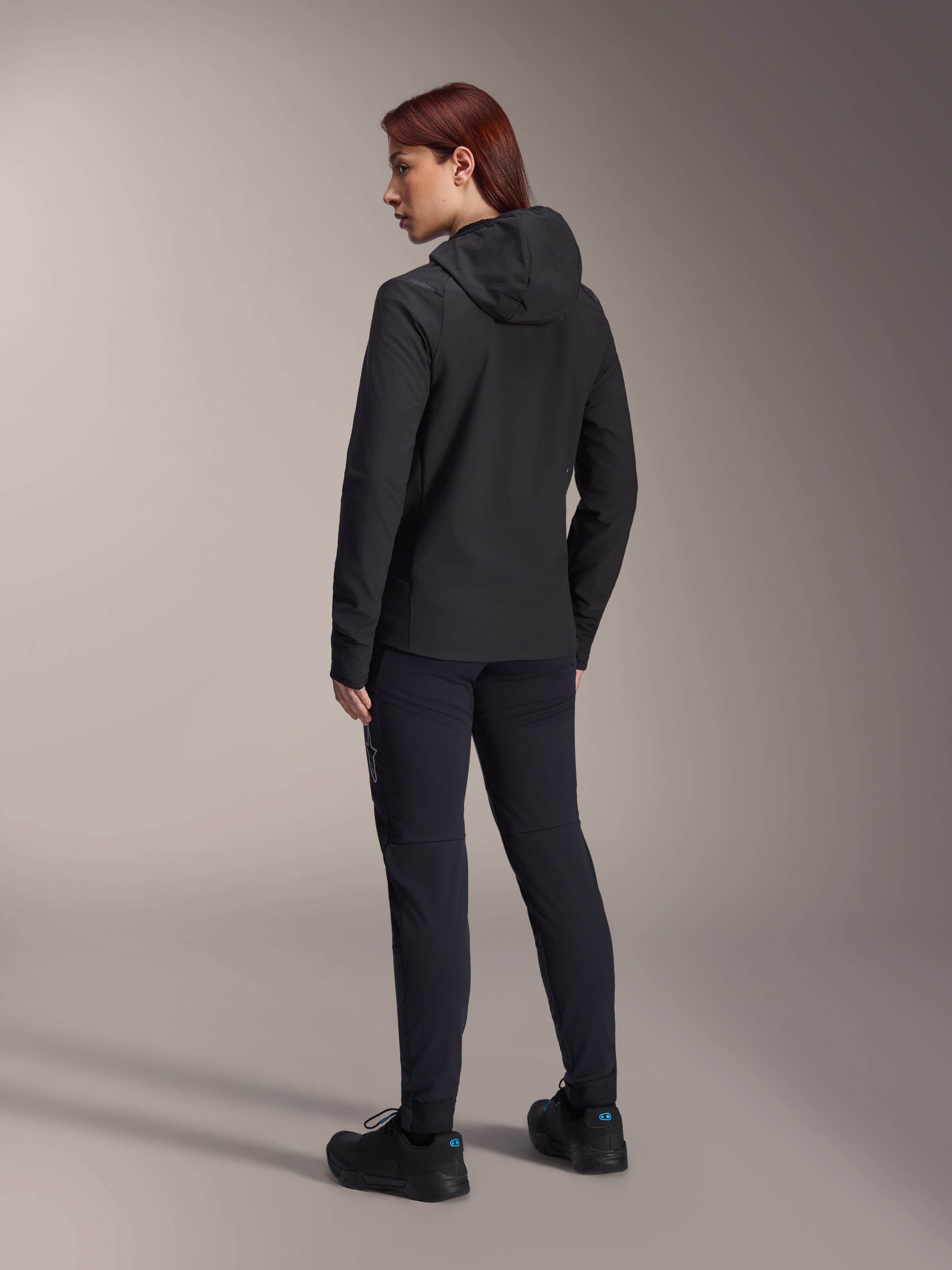 Alpinestars Stella A-Dura termojakke, MTB-cykeljakke til kvinder, sort, set bagfra med fokus på den isolerede hætte og regulær pasform, båret med matchende sorte bukser og sko, letvægts termisk isolering til kørsel i koldt vejr
