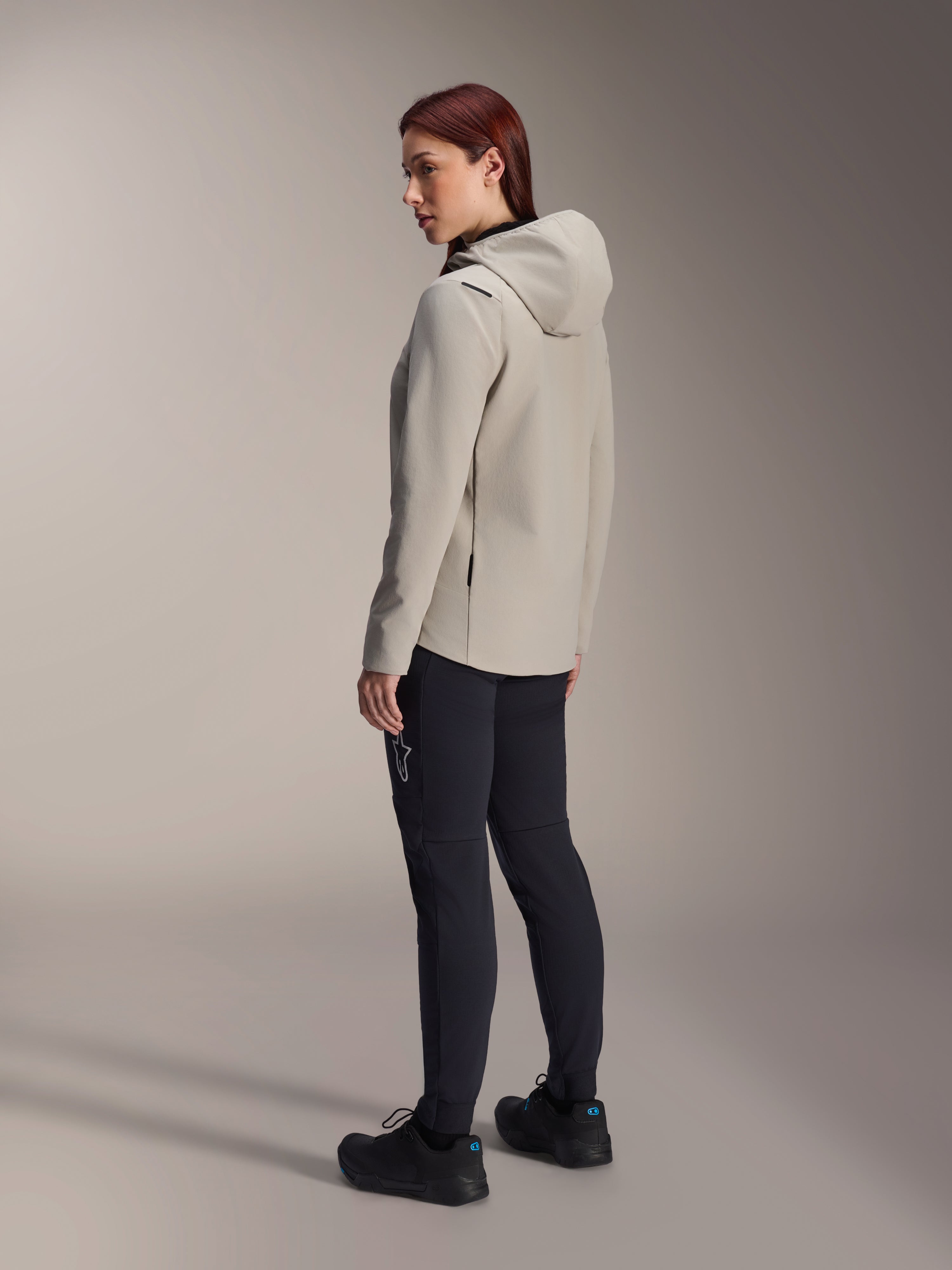Alpinestars Stella A-Dura Thermal Jacket i Titanium Gray, trekvart visning bagfra af en kvinde iført den isolerede cykeljakke med hætte og sorte MTB-bukser, studiebillede der viser den regulære pasform og lommedetaljer.