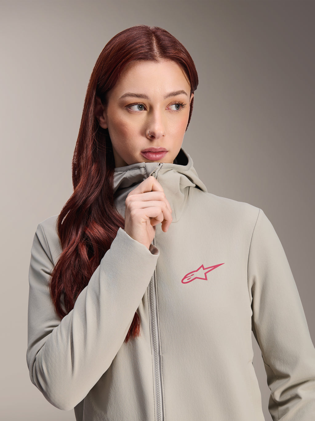 Alpinestars Stella A-Dura termojakke til kvinder, titaniumgrå med lyserødt Alpinestars-stjernelogo, model med langt rødt hår, der bærer og lyner den hætteklædte jakke, letvægtsisoleret cykeltøj til koldt vejr