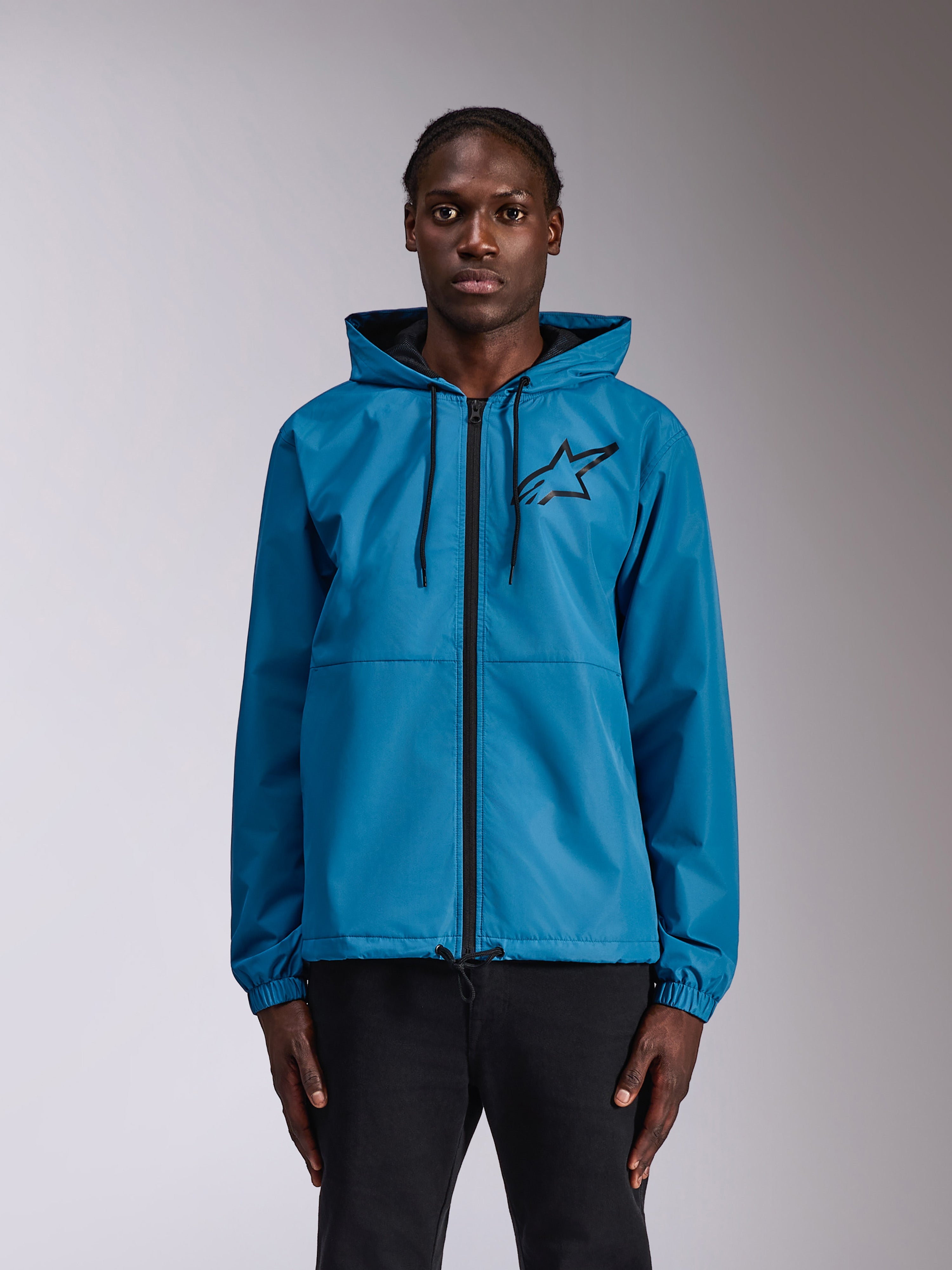 Alpinestars Speeder Chromium Windbreaker Jakke, blå vindjakke med hætte og sort Astars-logo på brystet, gennemgående lynlås foran, elastiske manchetter og hætte med snøre, båret af en model som afslappet sportstøj.