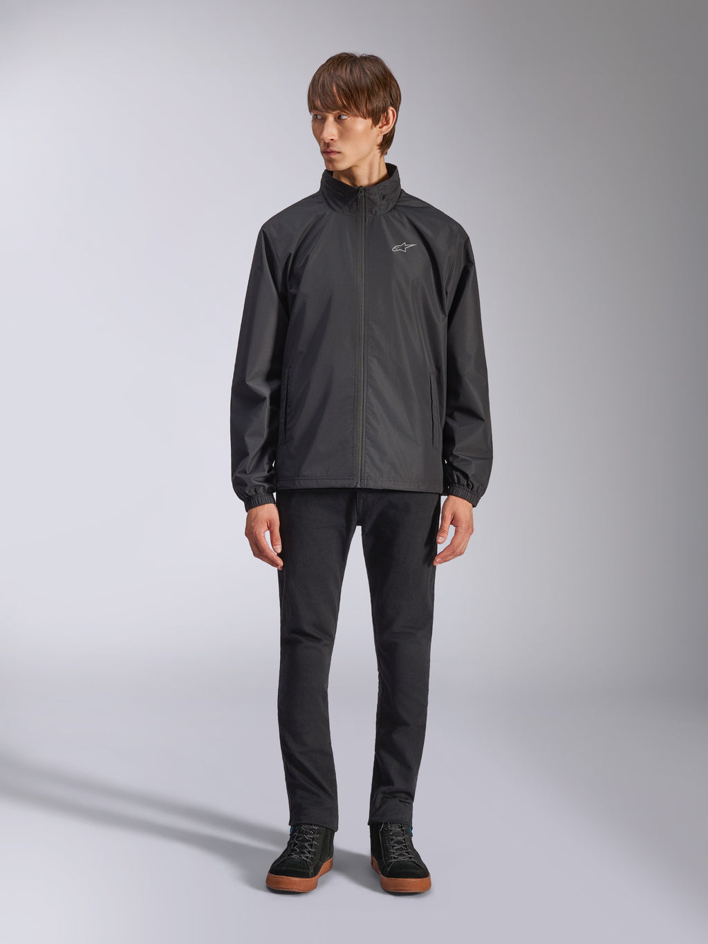 Alpinestars Overhaul Racer Jacket, Black Men's Rain Shell, full-length view on a model paired with black pants and casual sneakers, featuring a small white logo on the chest, water-resistant fabric, and casual motorsport-inspired design
