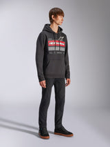 Alpinestars Harken hættetrøje, sort pullover fleece-hættetrøje, med stor grafik på brystet med rødt Alpinestars-navnetræk og stjernelogo, kængurulomme, båret af en model med sorte bukser og sneakers, afslappet motorsport-streetwear