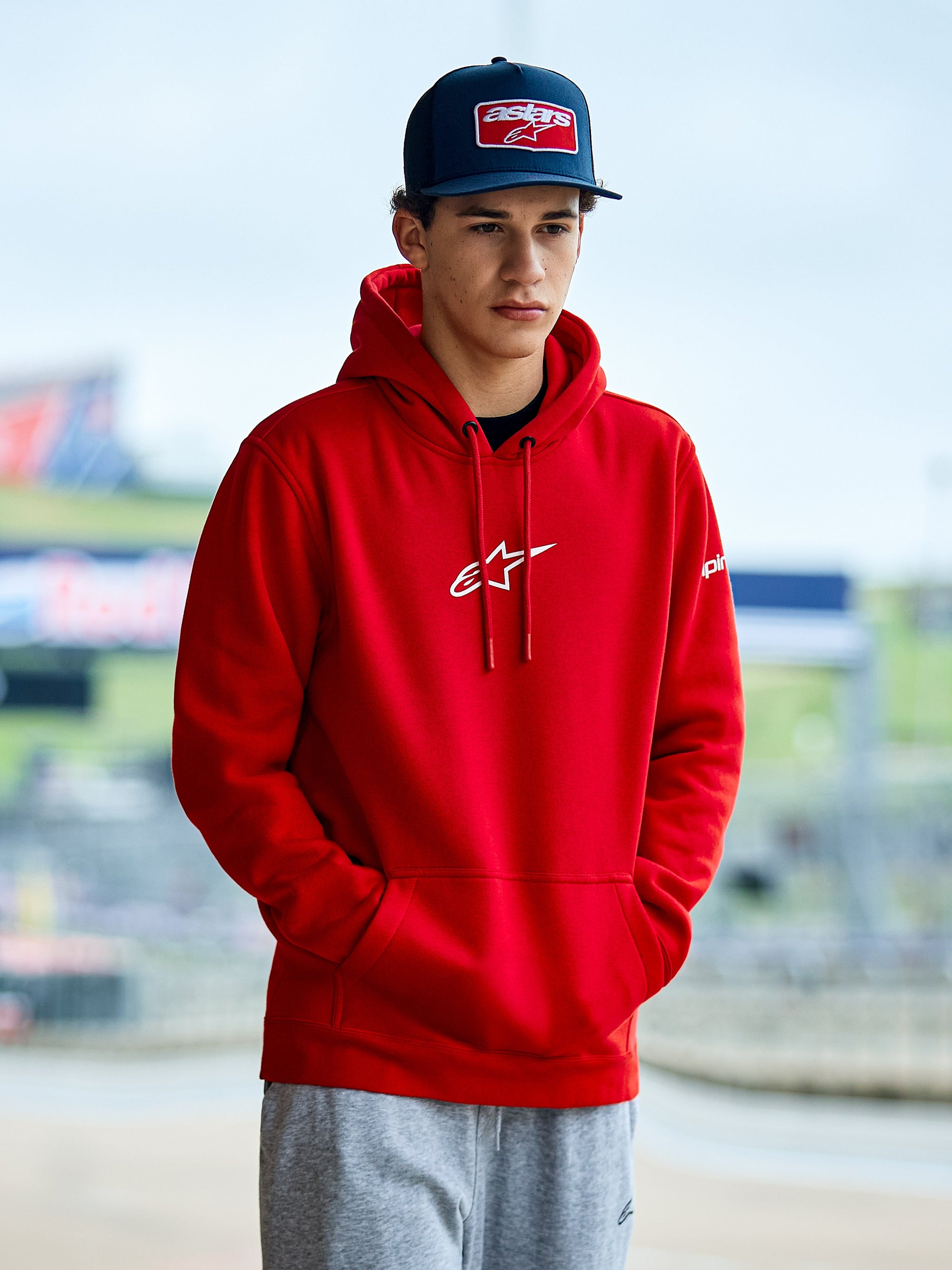 Alpinestars Frontal hættetrøje, rød pullover fleece hættetrøje, med hvidt Astars-logo på brystet, kængurulomme og hætte med snoretræk, båret af en mand med blå kasket, afslappet motorsport streetwear
