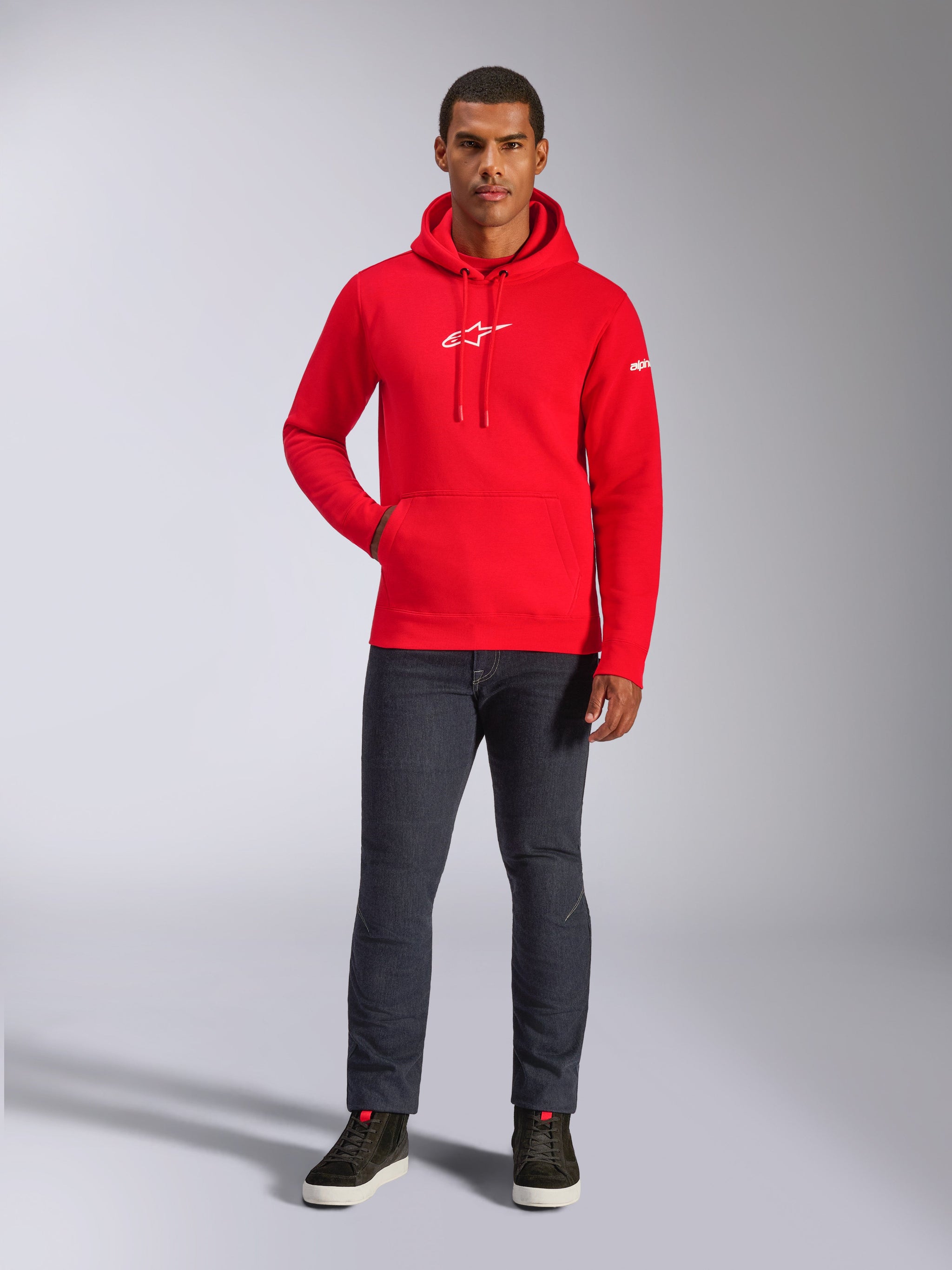 Mandlig model iført Alpinestars Frontal hættetrøje i rød, pullover fleece-hættetrøje med hvidt Astars-logo på brystet, kængurulomme, hætte med snoretræk, kombineret med mørkegrå jeans og sorte high-top sneakers, casual motorsport-streetwear