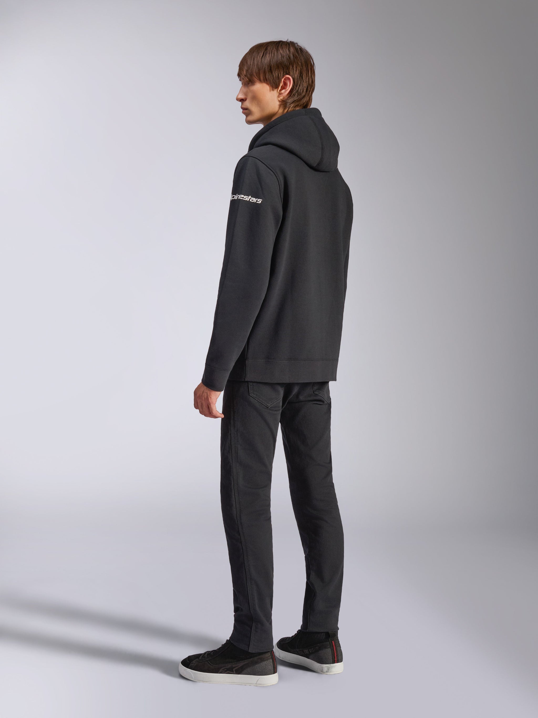 Alpinestars Frontal hættetrøje, afslappet streetwear, sort, set skråt bagfra, viser hvidt ordmærke-logo på ærmet, båret med sorte bukser og sneakers, moderne motorsport-inspireret design