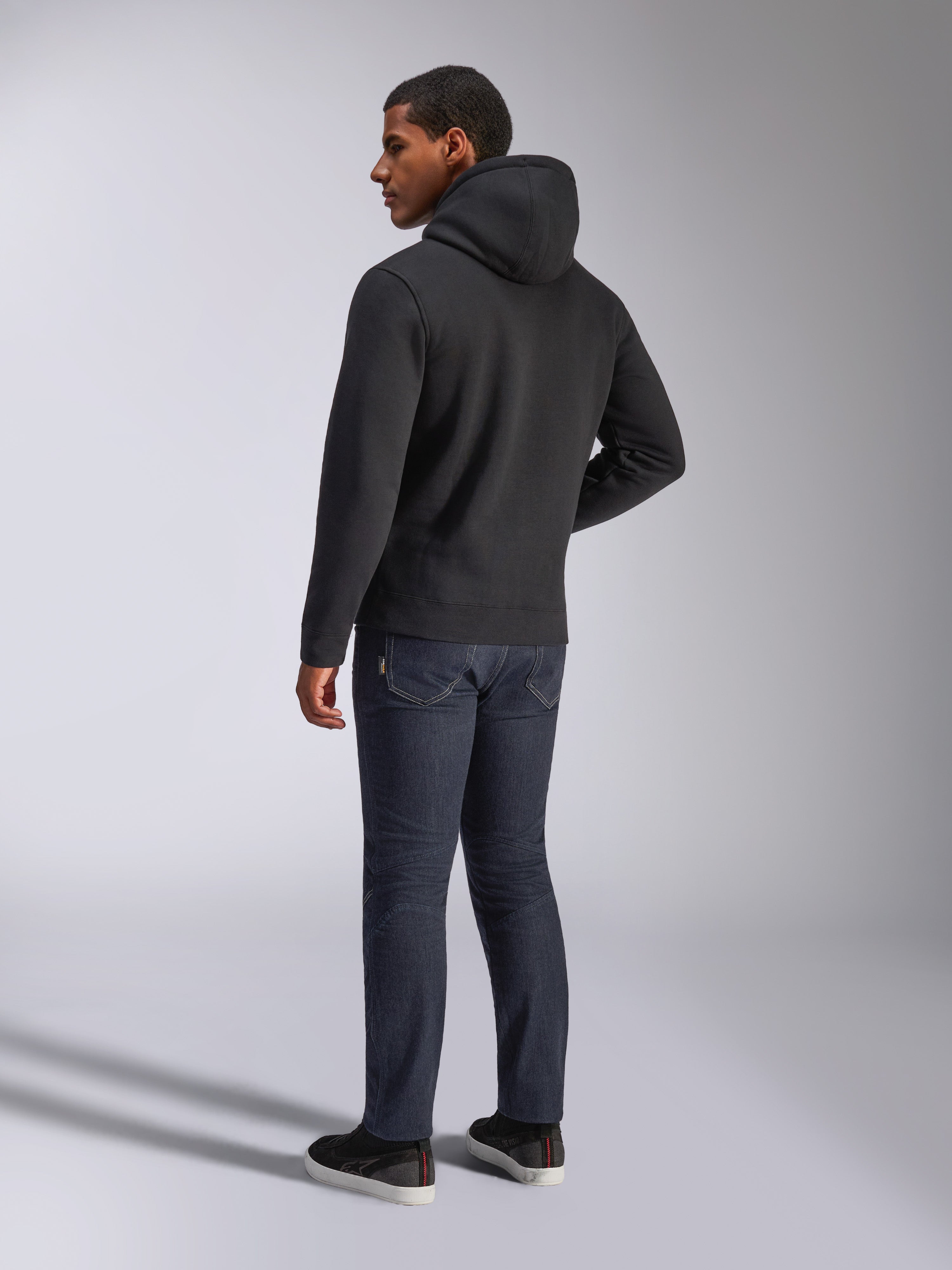 Alpinestars Blaze V3 hættetrøje, set bagfra på en mand iført den sorte pullover-hættetrøje, kombineret med mørke jeans og sorte sneakers, afslappet motorsport-inspireret streetwear