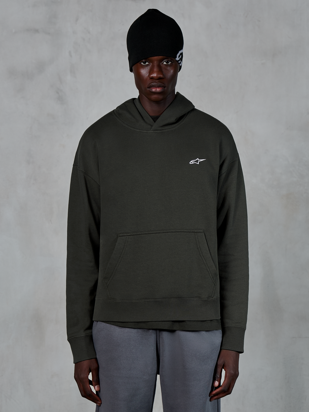 Alpinestars Essentials Ageless Hoodie, casual streetwear, Off Black pullover med et lille hvidt Alpinestars-logo på brystet, båret med en sort beanie, minimalistisk design med kængurulomme og stenvasket finish