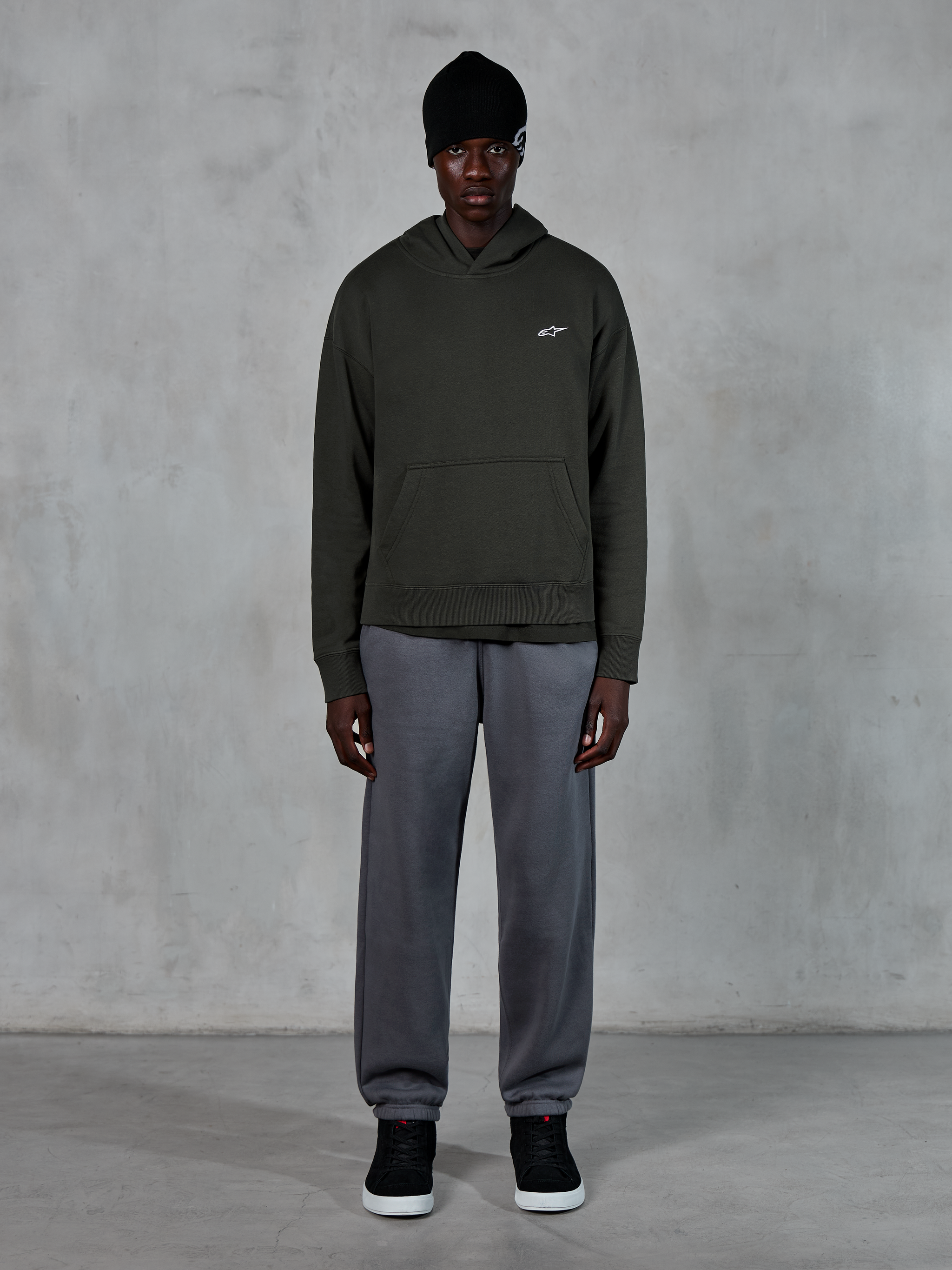 Model wearing Alpinestars Essentials Ageless Hoodie, casual streetwear, Off Black with small white logo on chest, paired with gray joggers, black beanie, and black sneakers, minimalist motorsport-inspired design