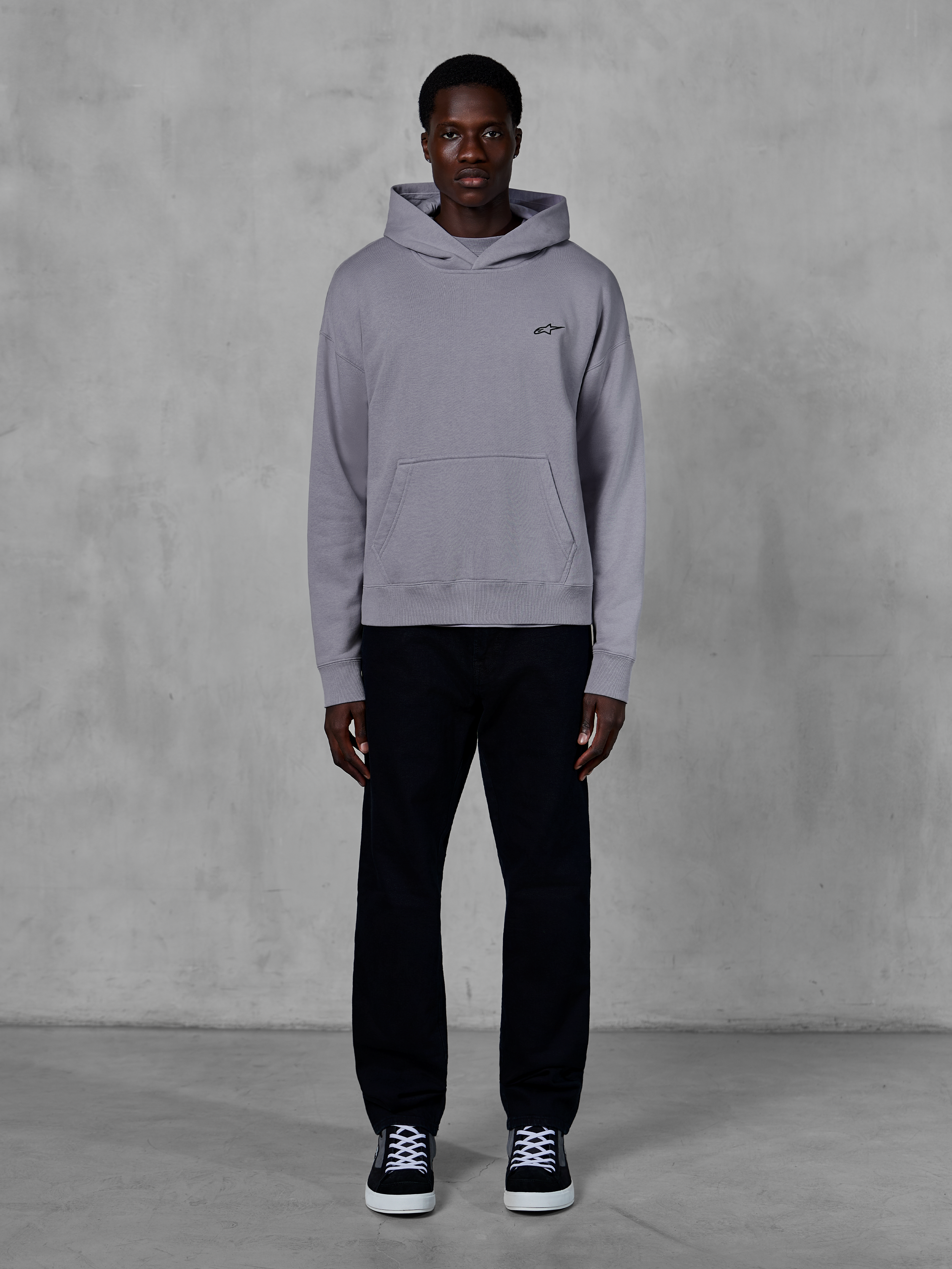 Alpinestars Essentials Ageless Hoodie, Silver, mandlig model i afslappet streetwear, lille sort Alpinestars-logo på brystet, kængurulomme foran, minimalistisk design, kombineret med sorte bukser og sneakers.