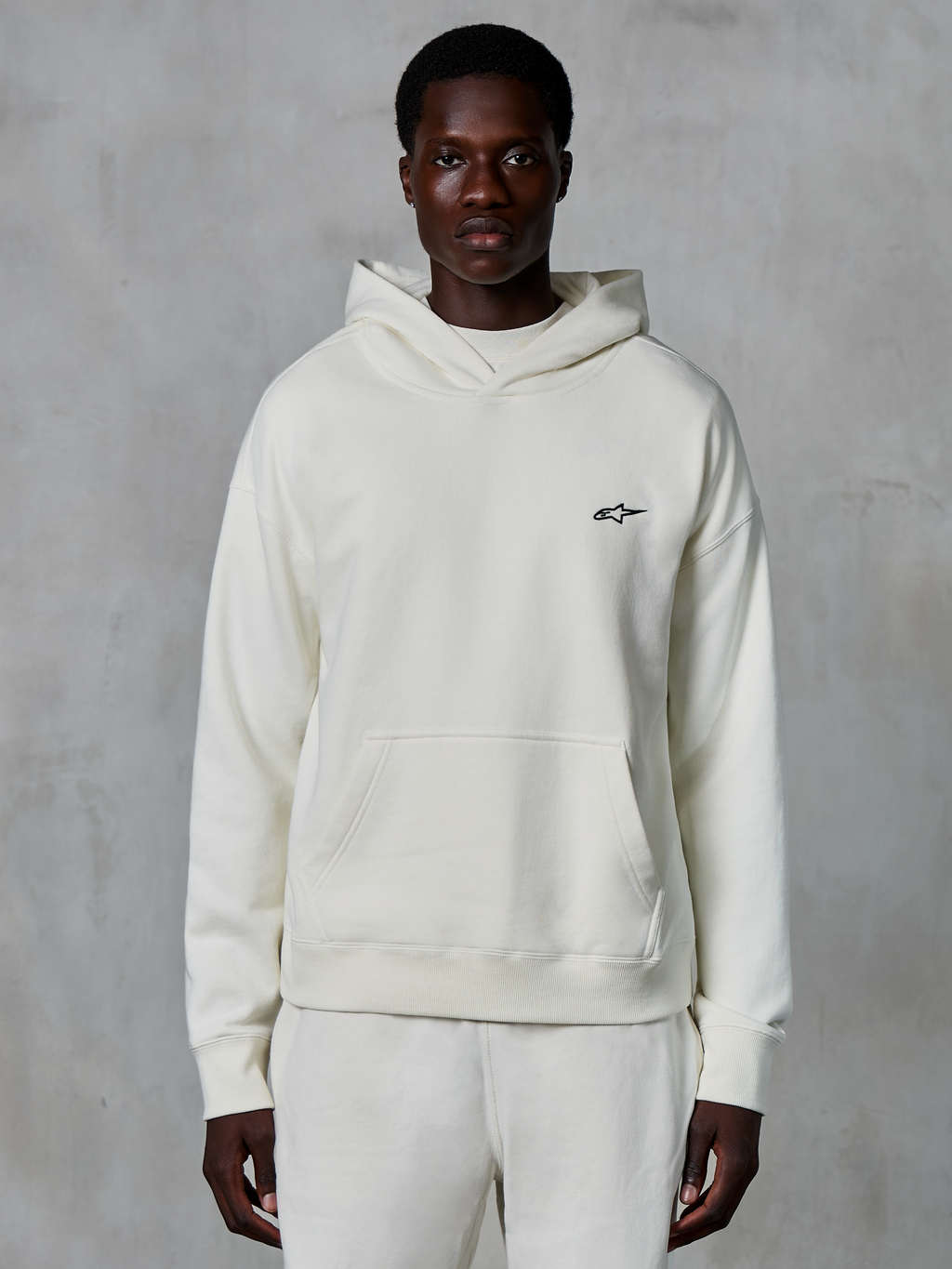 Alpinestars Essentials Ageless hættetrøje, off-white, båret af en mandlig model, lille sort logo på brystet, kængurulomme foran, lange ærmer, afslappet motorsport-inspireret streetwear
