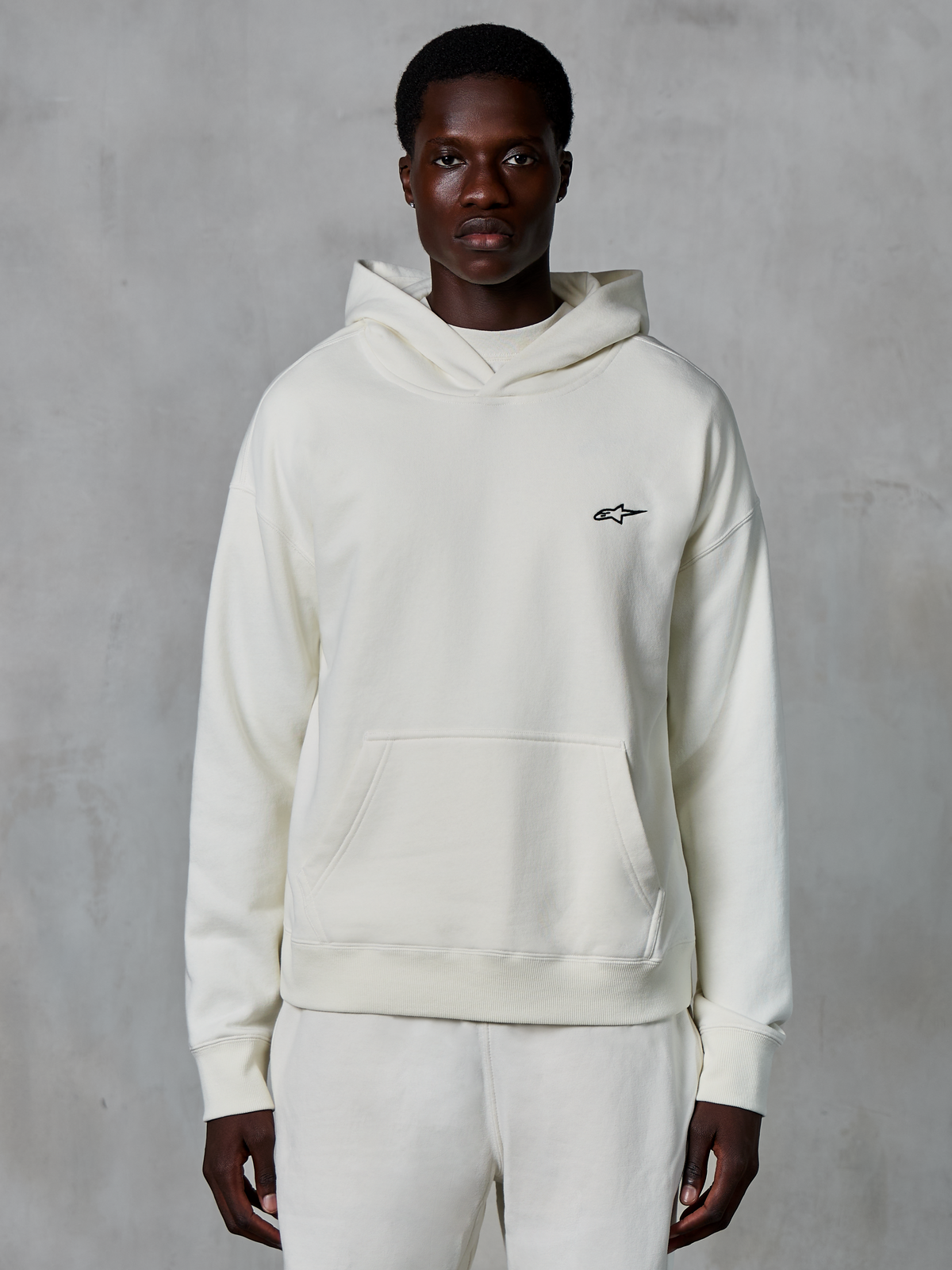Alpinestars Essentials Ageless hættetrøje, off-white, båret af en mandlig model, lille sort logo på brystet, kængurulomme foran, lange ærmer, afslappet motorsport-inspireret streetwear