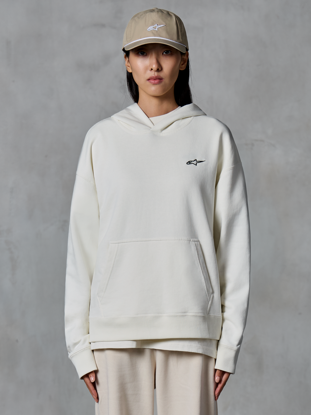 Alpinestars Essentials Ageless hættetrøje, off-white pullover fleece-hættetrøje, kvindelig model iført sandfarvet kasket og matchende creme-farvede joggingbukser, lille sort Alpinestars-logo på brystet, kængurulomme foran, afslappet pasform, afslappet motorsport-inspireret streetwear