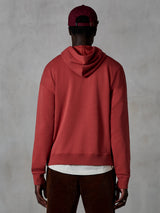 Alpinestars Essentials Ageless Hoodie, afslappet fleece-hættetrøje, teglrød, set bagfra af model iført livsstilstøj, enkelt design med ribkant forneden og ved manchetterne, stylet med brune fløjlsbukser og en bordeaux kasket