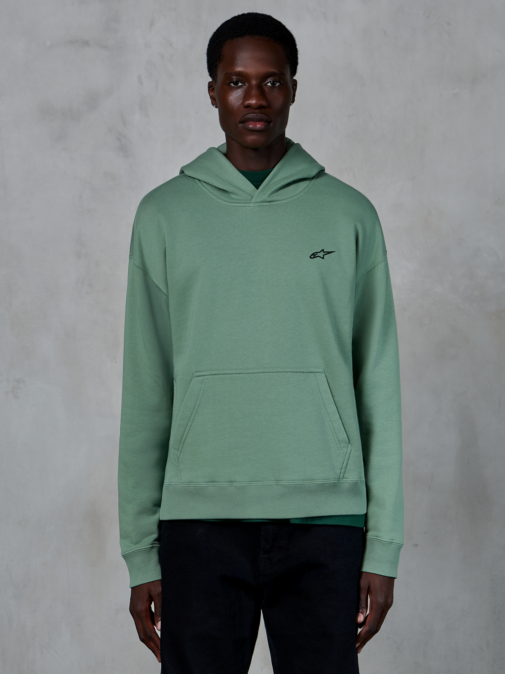 Alpinestars Essentials Ageless Hoodie, kaktusgrøn, afslappet fleece-hættetrøje med stenvasket finish, lille sort stjernelogo på brystet, kængurulomme, båret af en model i et stilrent livsstilsmiljø
