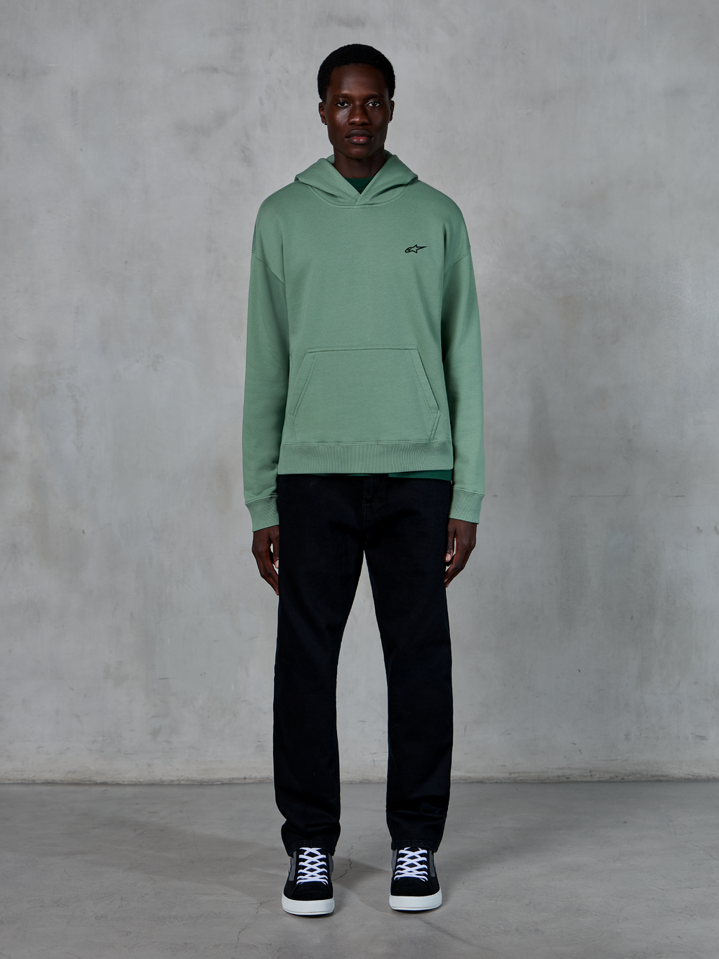 Alpinestars Essentials Ageless hættetrøje, kaktusgrøn, lille sort logo på brystet, kængurulomme, afslappet lifestyle streetwear kombineret med sorte bukser og sort-hvide sneakers mod en tekstureret væg.