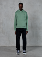 Alpinestars Essentials Ageless hættetrøje, kaktusgrøn, lille sort logo på brystet, kængurulomme, afslappet lifestyle streetwear kombineret med sorte bukser og sort-hvide sneakers mod en tekstureret væg.