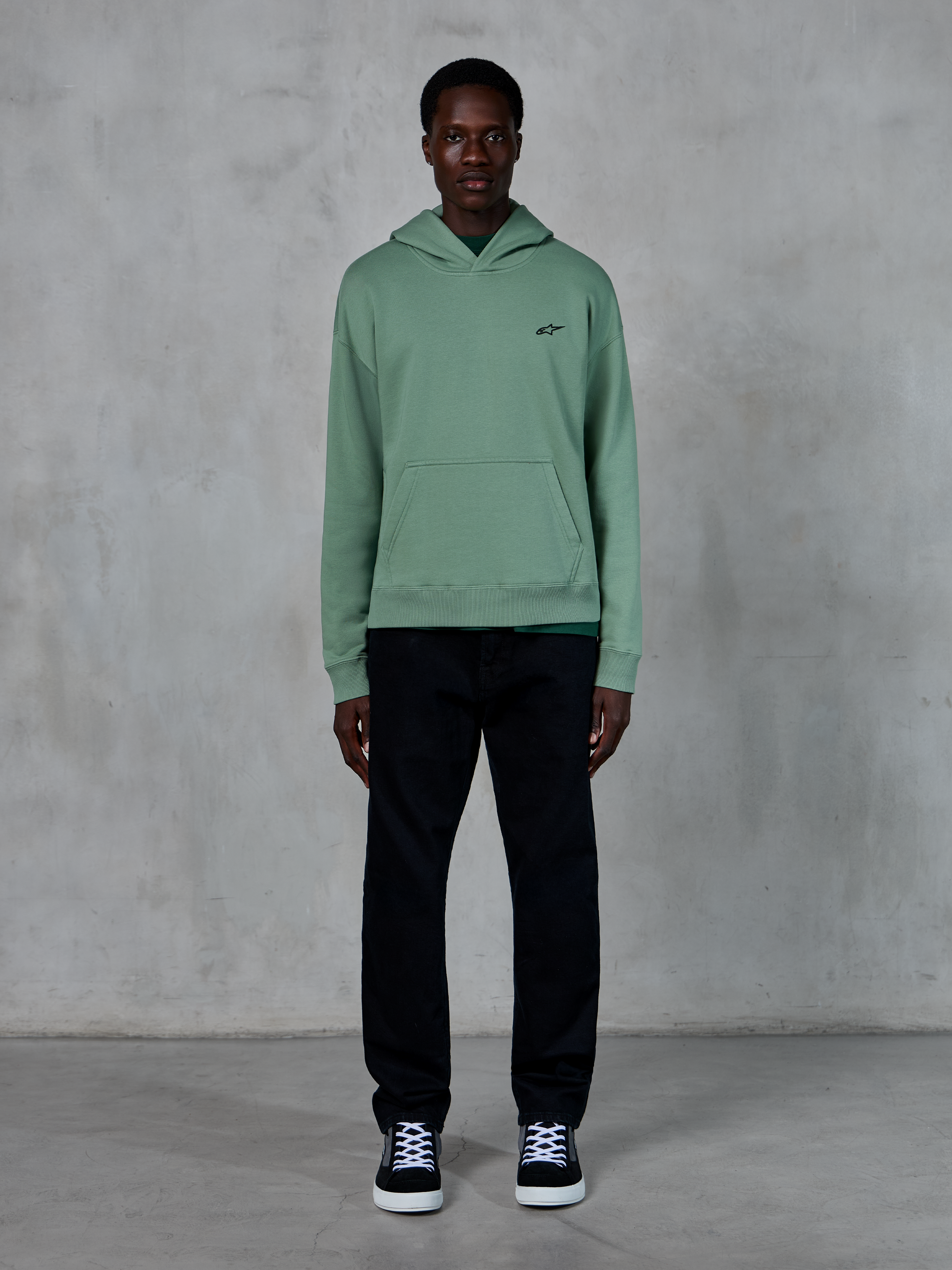 Alpinestars Essentials Ageless hættetrøje, kaktusgrøn, lille sort logo på brystet, kængurulomme, afslappet lifestyle streetwear kombineret med sorte bukser og sort-hvide sneakers mod en tekstureret væg.