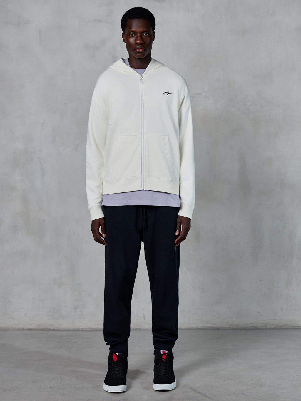 Alpinestars Essentials Ageless Zip Hoodie, off-white hættetrøje med gennemgående lynlås, afslappet pasform med lille sort logo på brystet, båret med sorte joggingbukser og sneakers, afslappet motorsport-inspireret streetwear.