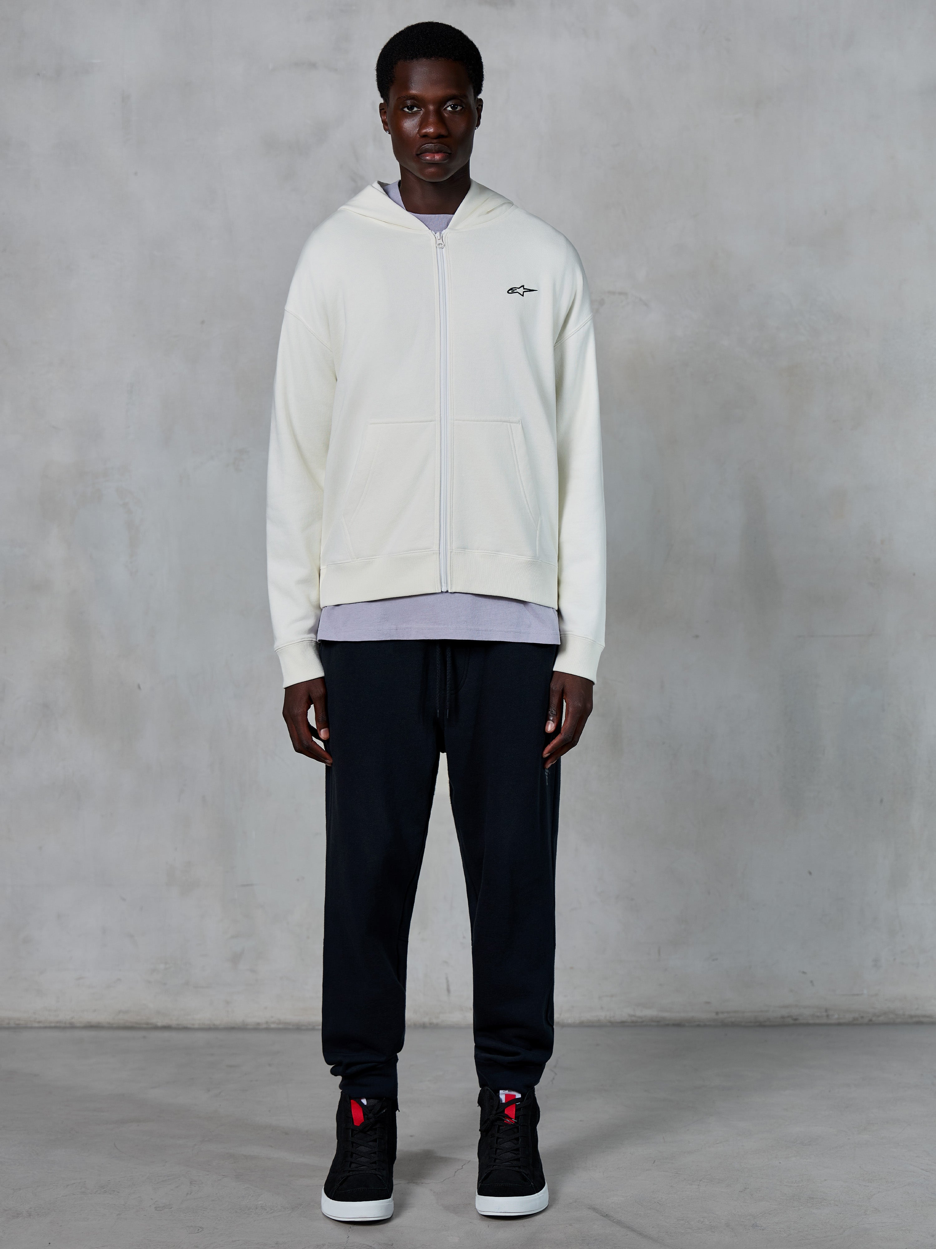 Alpinestars Essentials Ageless Zip Hoodie, off-white hættetrøje med gennemgående lynlås, afslappet pasform med lille sort logo på brystet, båret med sorte joggingbukser og sneakers, afslappet motorsport-inspireret streetwear.