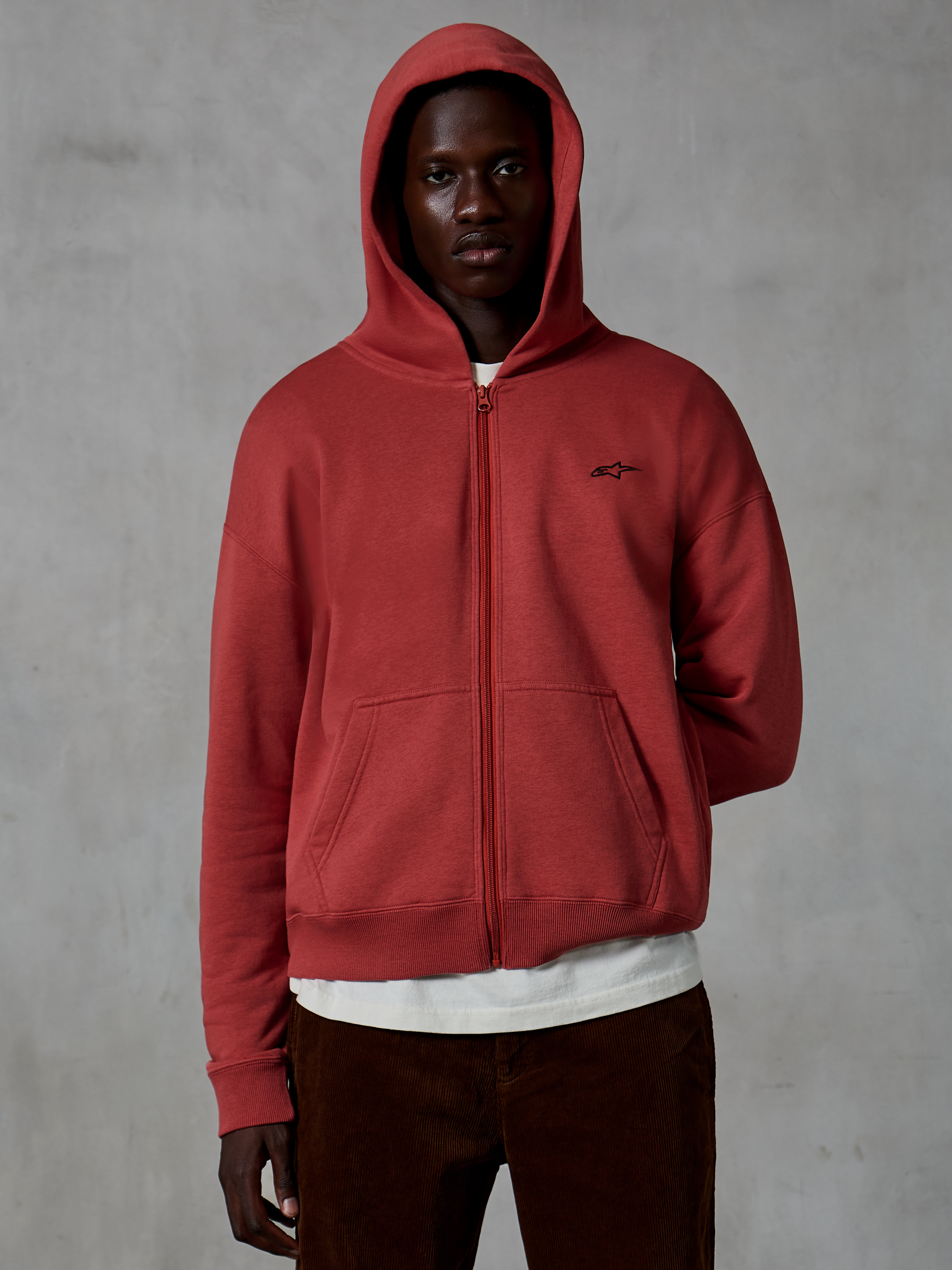 Alpinestars Essentials Ageless Zip Hoodie, afslappet sportswear i Brick rød, design med gennemgående lynlås og hætten oppe, lille sort logo på brystet, båret af en model med hvid t-shirt og brune fløjlsbukser mod en grå baggrund
